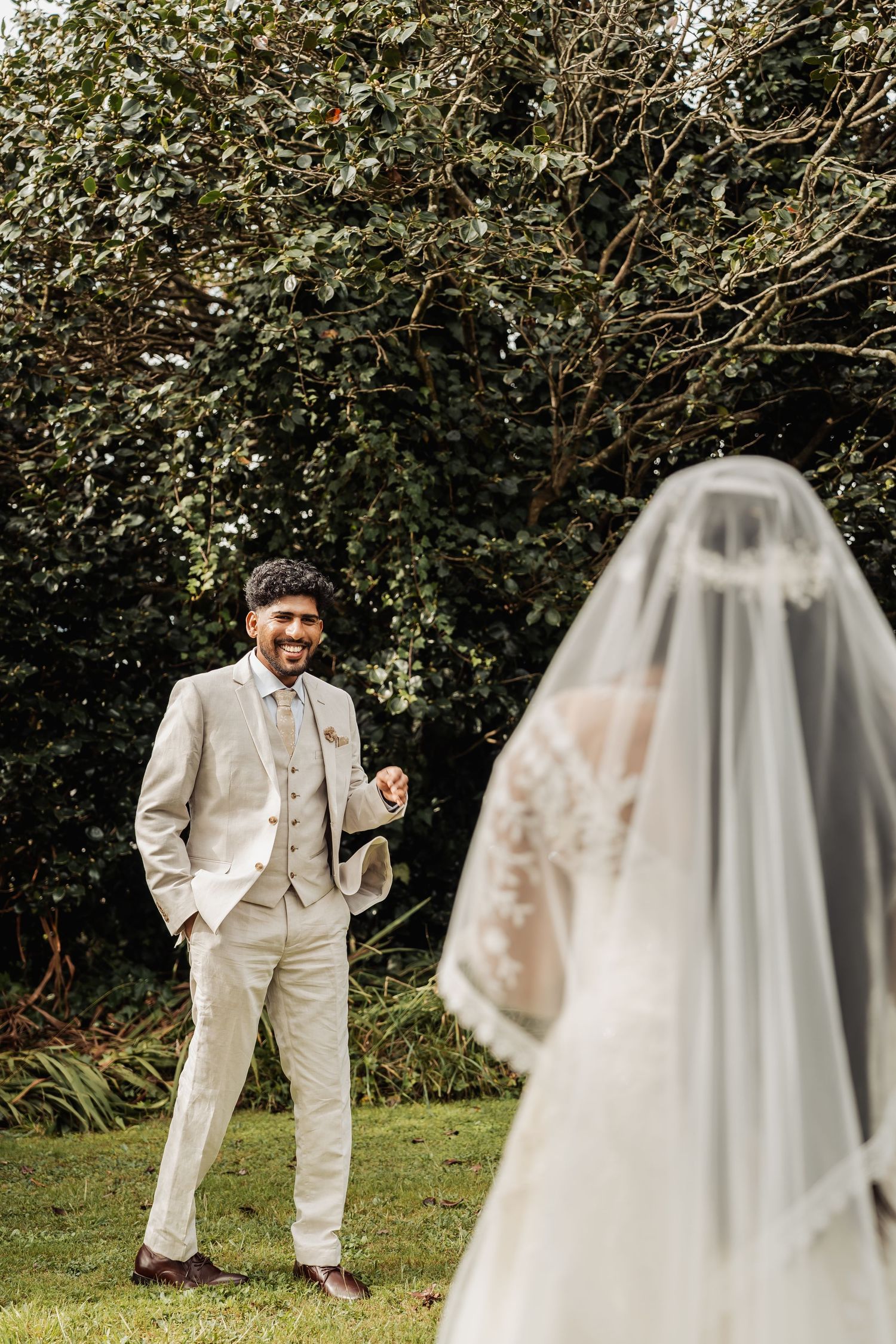 Smiling groom in beige suit reacts during first look with bride in white wedding dress and veil outdoors.