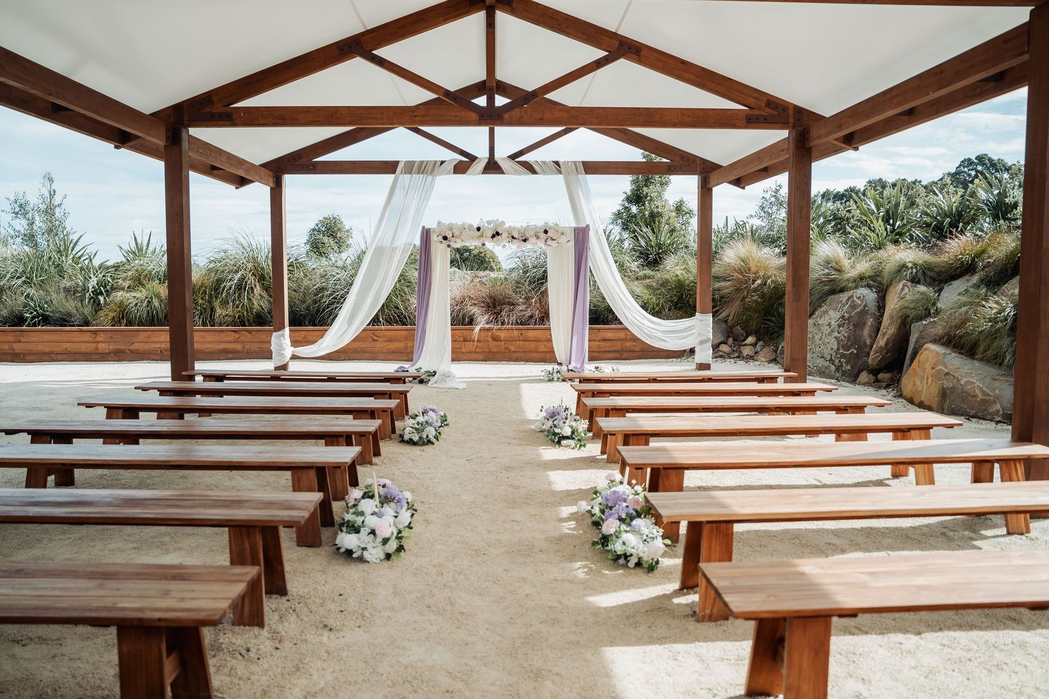 Elegant outdoor wedding ceremony setup with wooden pews, floral arrangements, and a draped arch under a rustic pergola.