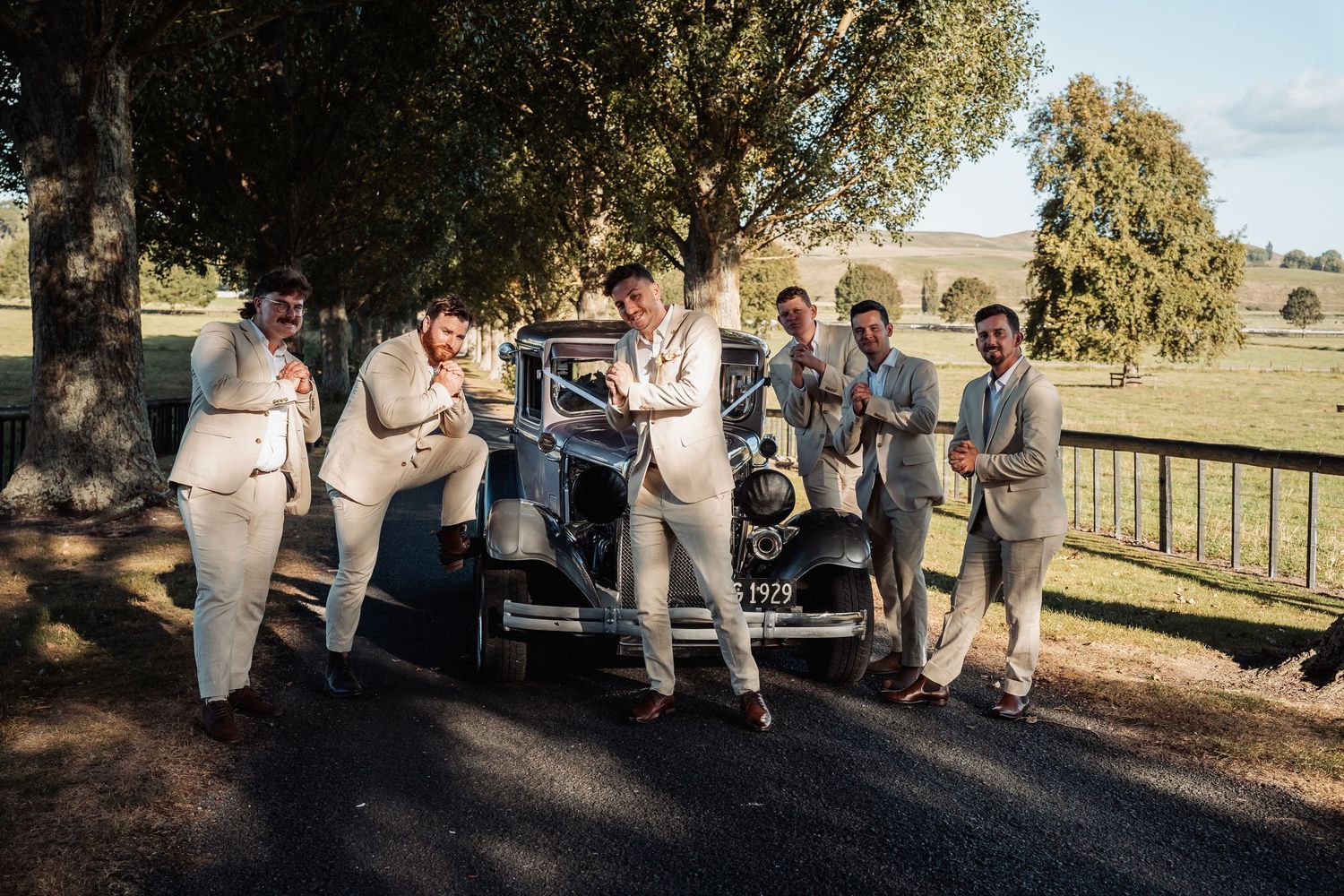 Groom and groomsmen in tan suits pose playfully beside vintage black car outdoors.