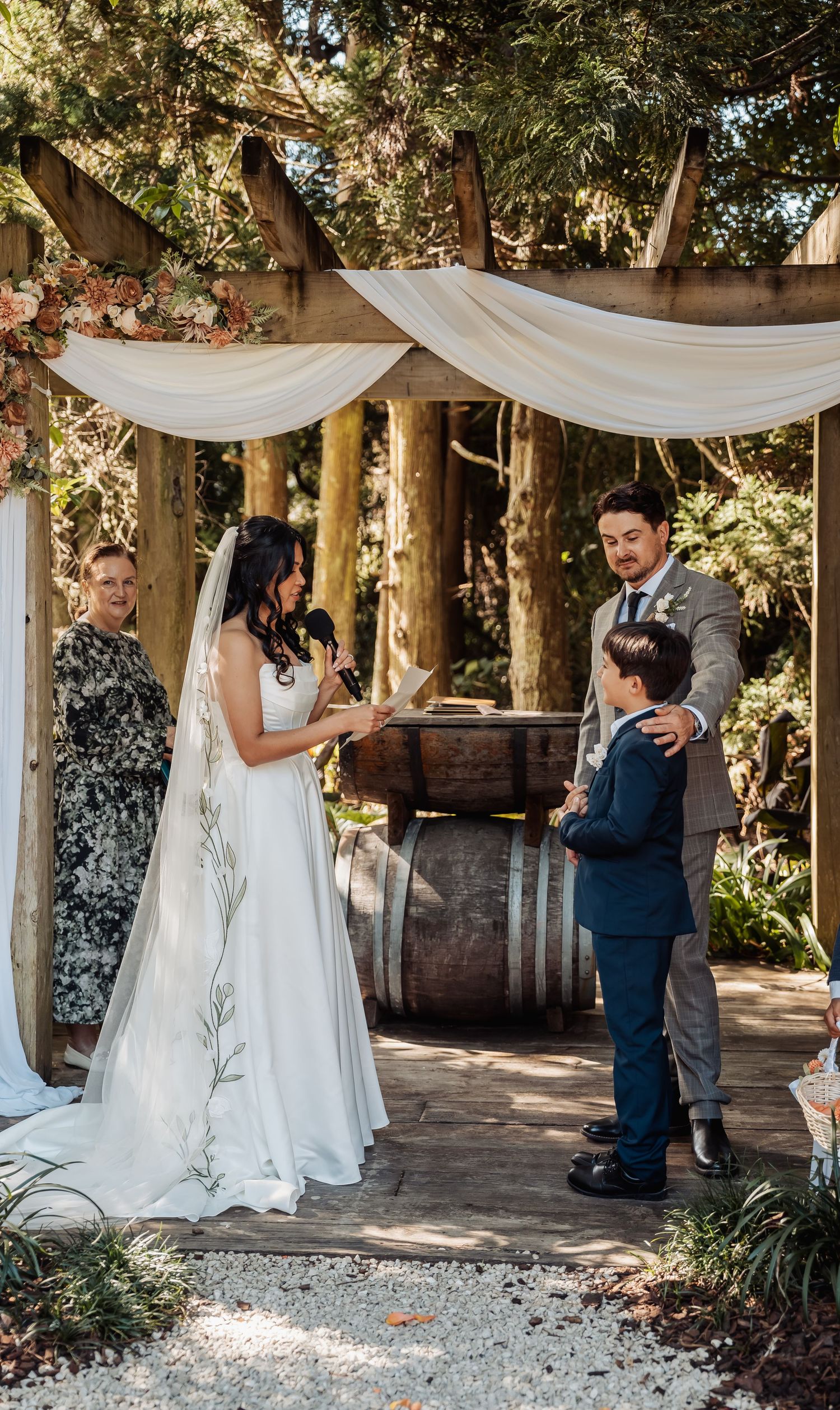 Bride reads vows to groom with young boy at outdoor woodland wedding ceremony under draped pergola.