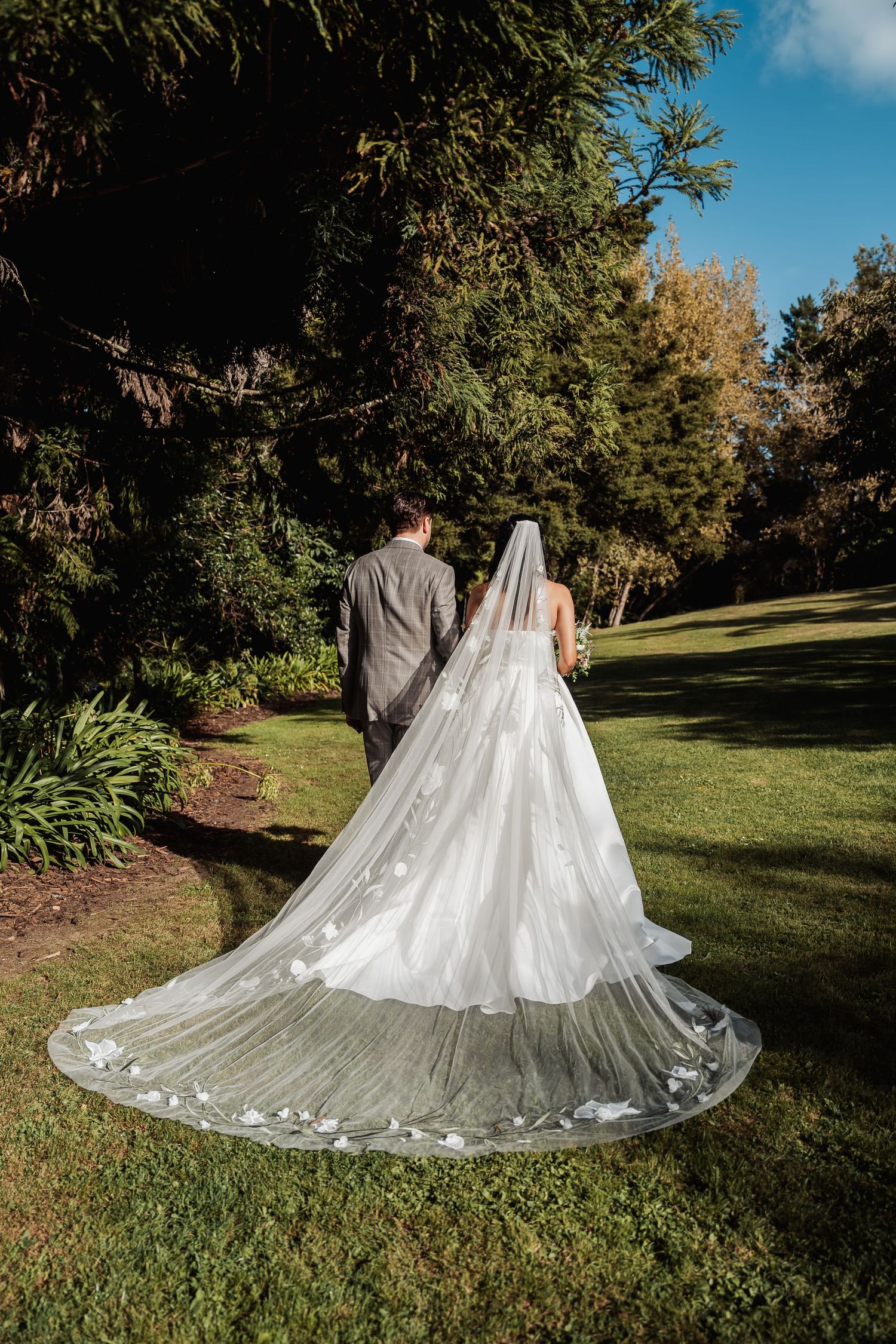 Bride and groom walk hand in hand through a lush garden, showcasing her stunning floral cathedral veil.