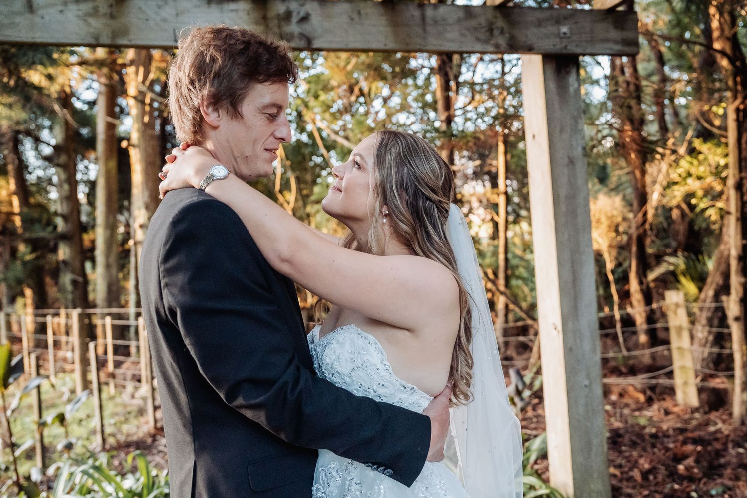 A bride in a white lace gown and groom in a black suit embrace under a wooden arbor during an outdoor autumn wedding.