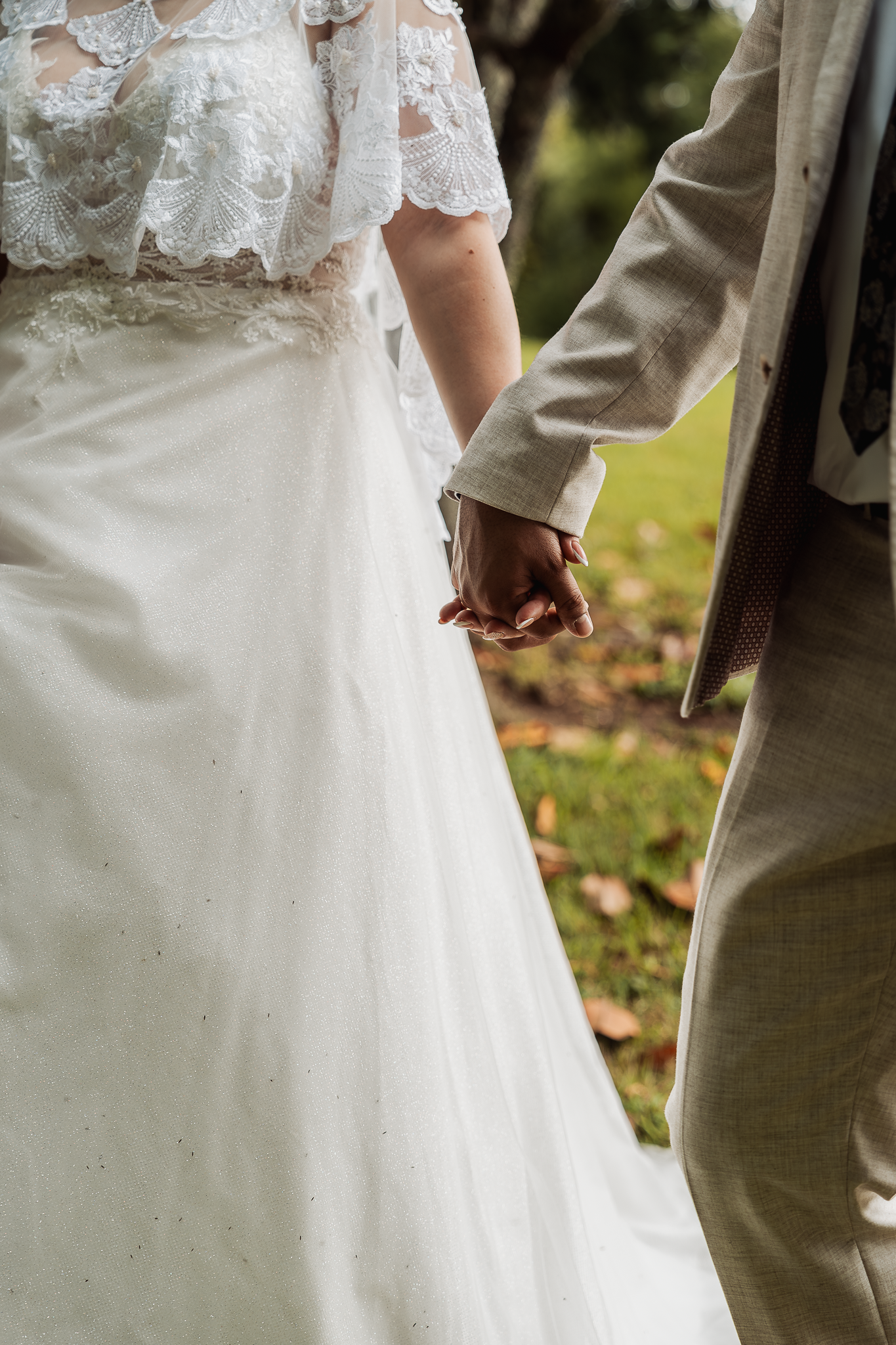 Close up of couple holding hands in wedding attire, showing lace dress detail and tan suit.