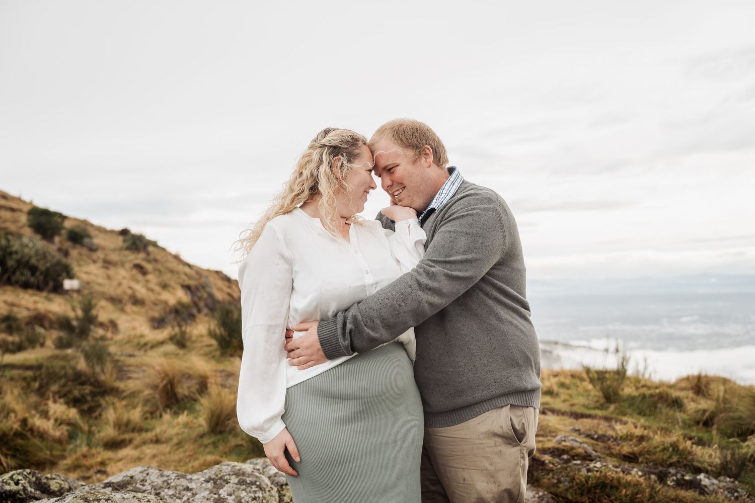 A romantic embrace on a windswept hilltop overlooking a misty valley on an overcast day.