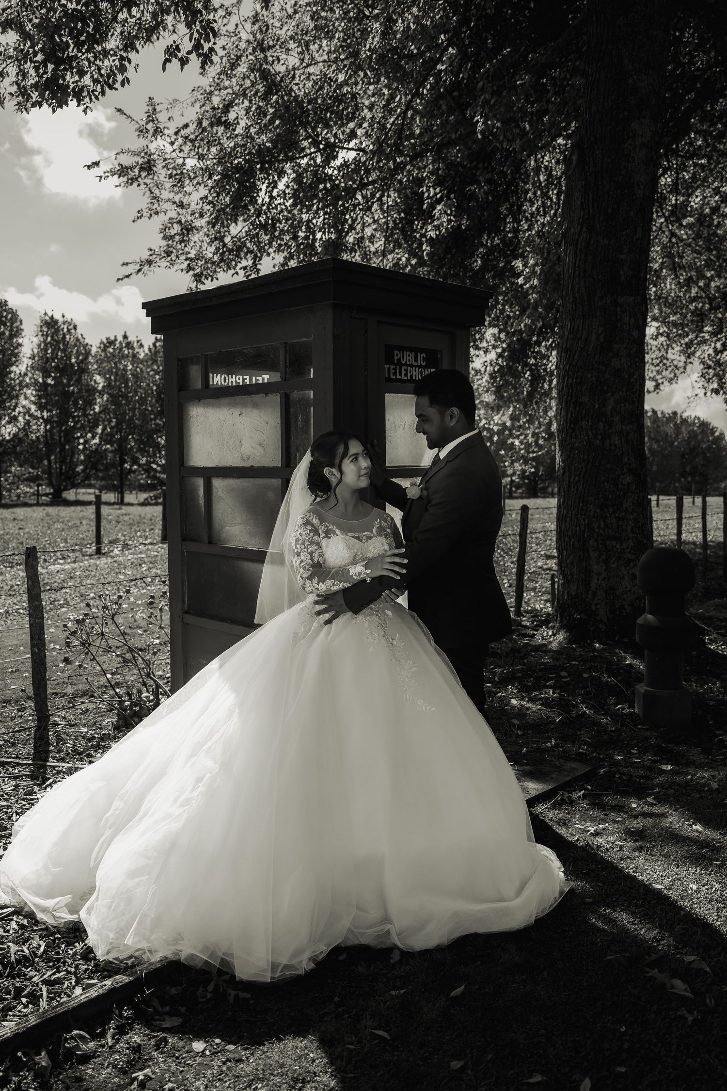 Black and white photo of bride and groom embracing beside a vintage phone booth under tall trees.