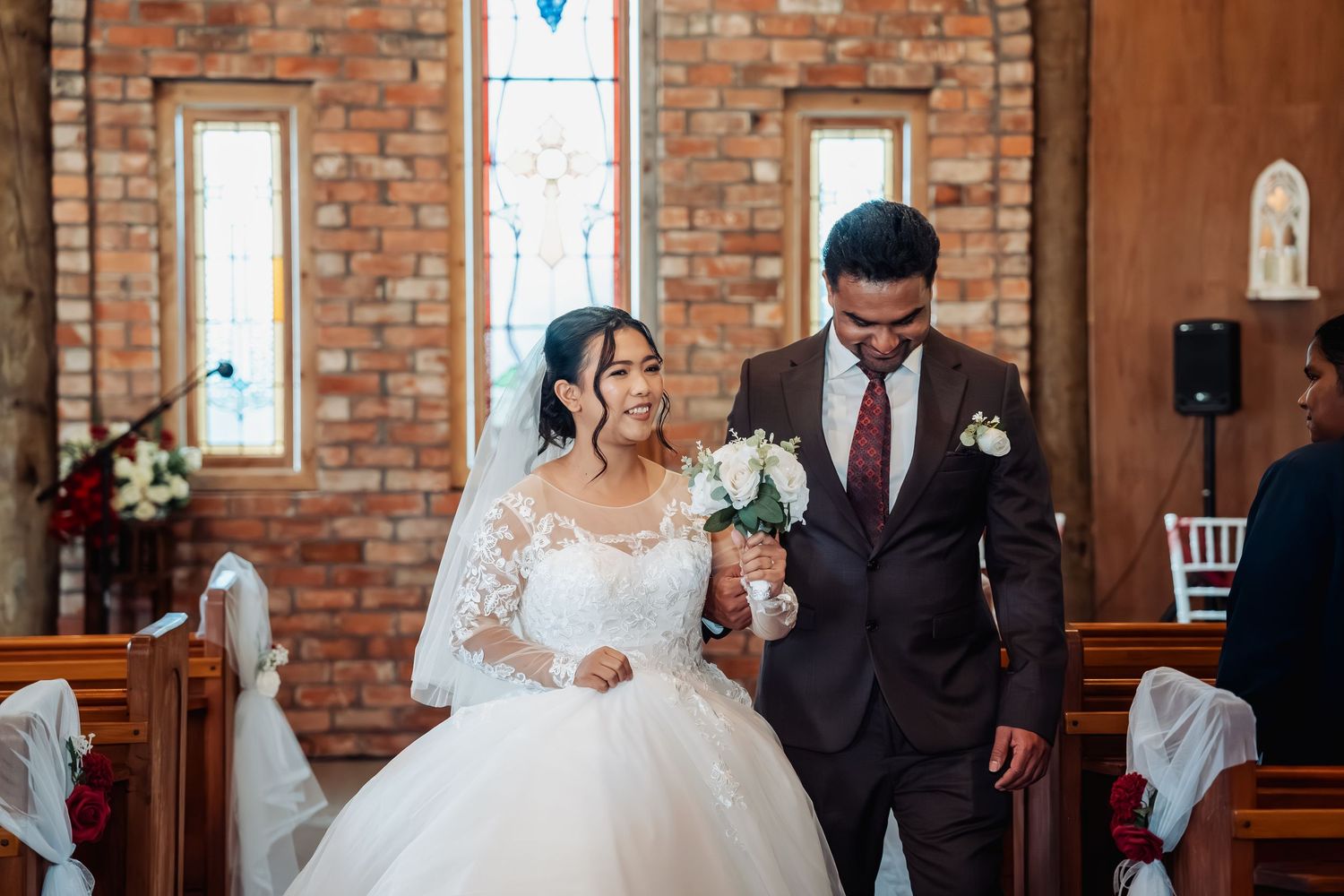Newlywed couple smiling joyfully while walking arm in arm through a charming brick-walled church after their ceremony.