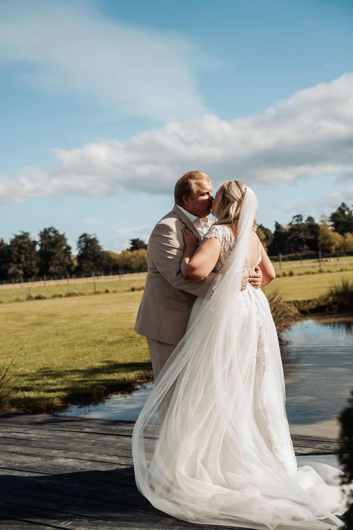 Bride and groom share a romantic embrace on a wooden deck overlooking a scenic green countryside on their wedding day.