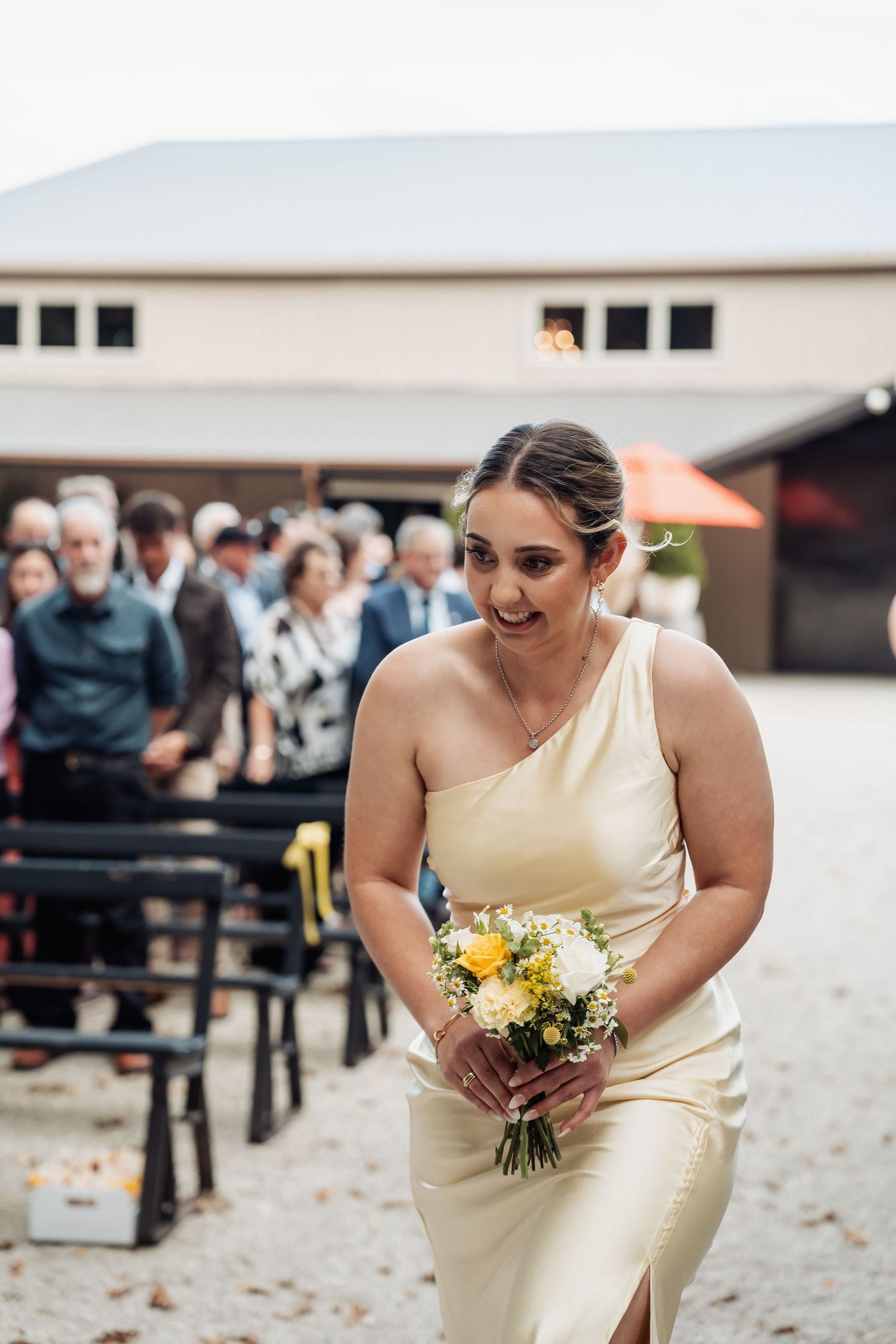 Bridesmaid in cream one-shoulder gown walks down outdoor wedding aisle holding yellow and white bouquet.