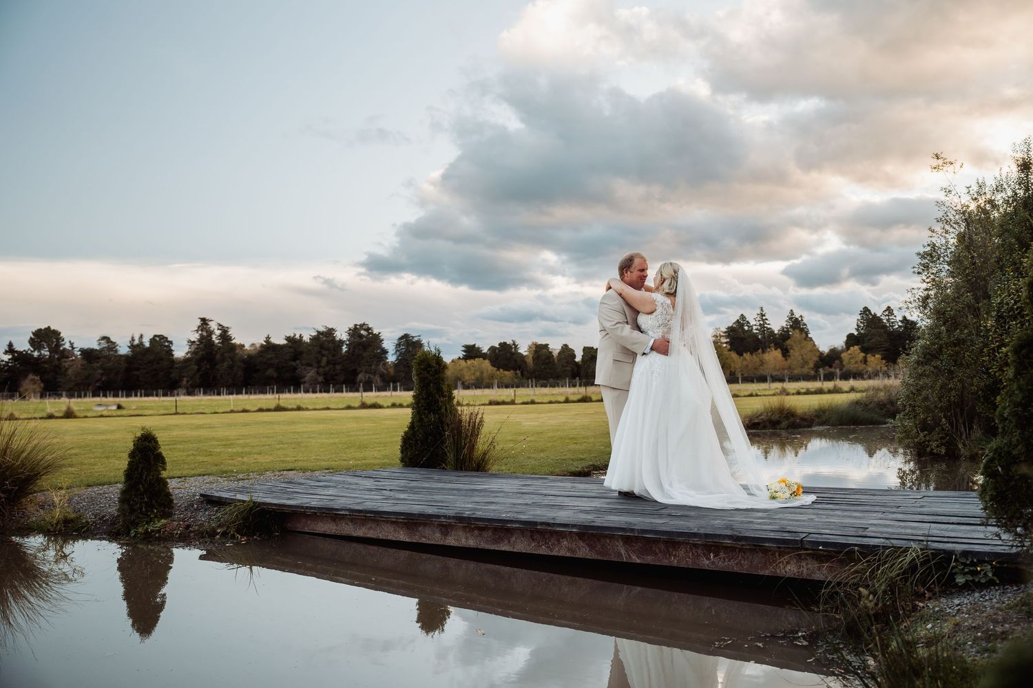 Bride and groom share a romantic kiss on a wooden dock overlooking a serene pond at sunset.