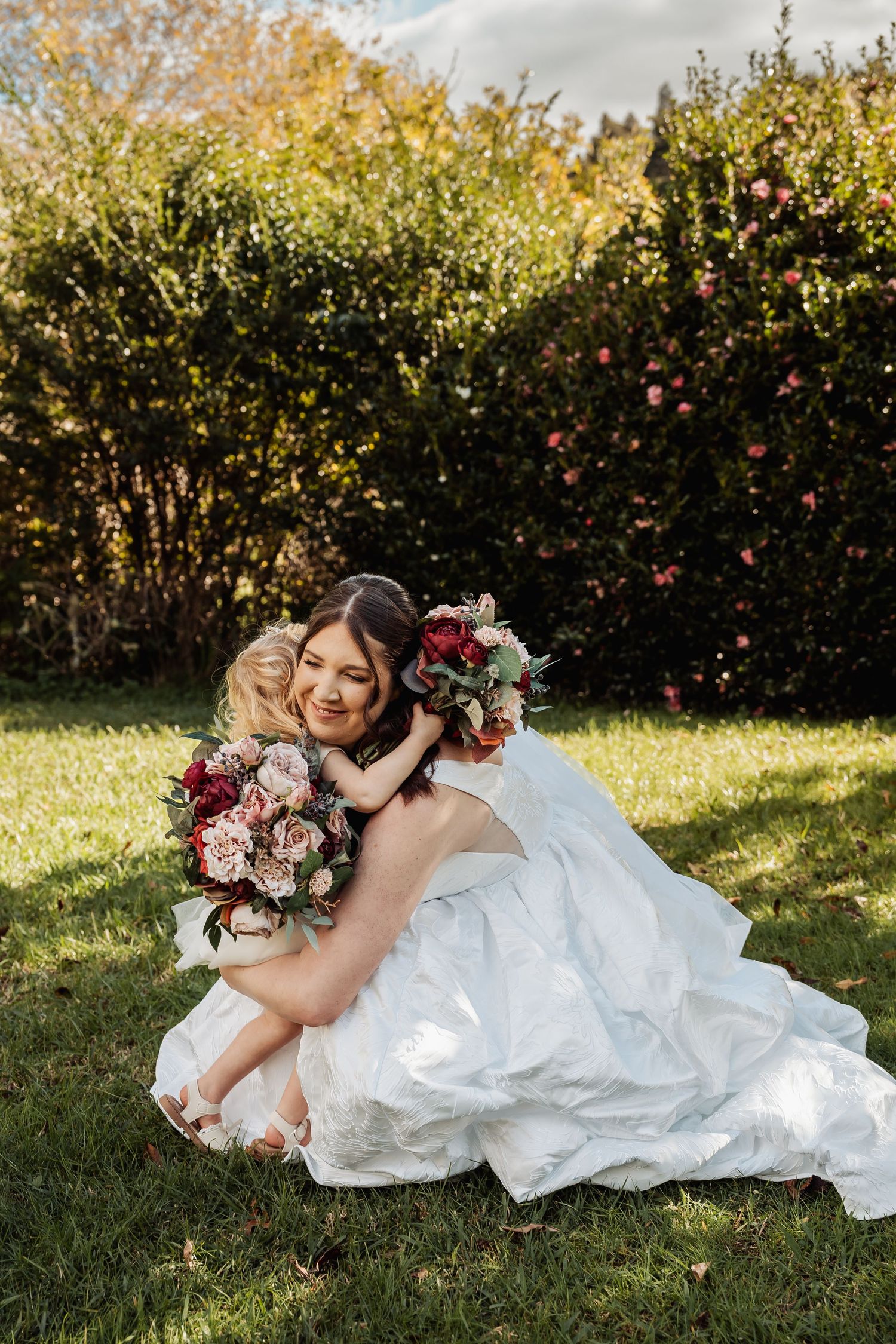 Smiling bride in white wedding gown hugs young flower girl while holding bouquet outdoors.