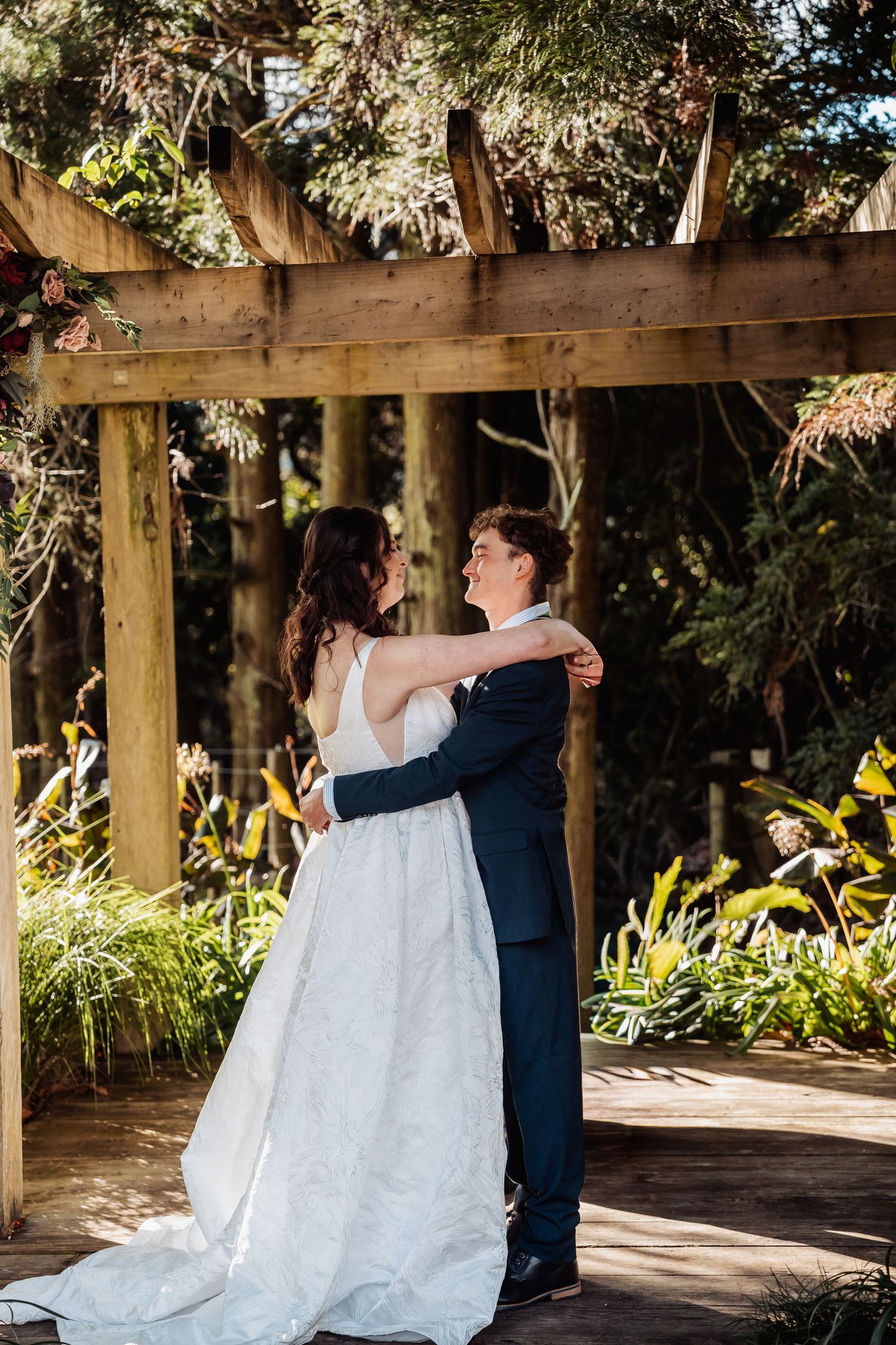 Bride and groom share a tender embrace under a wooden pergola surrounded by lush greenery at an outdoor wedding ceremony.