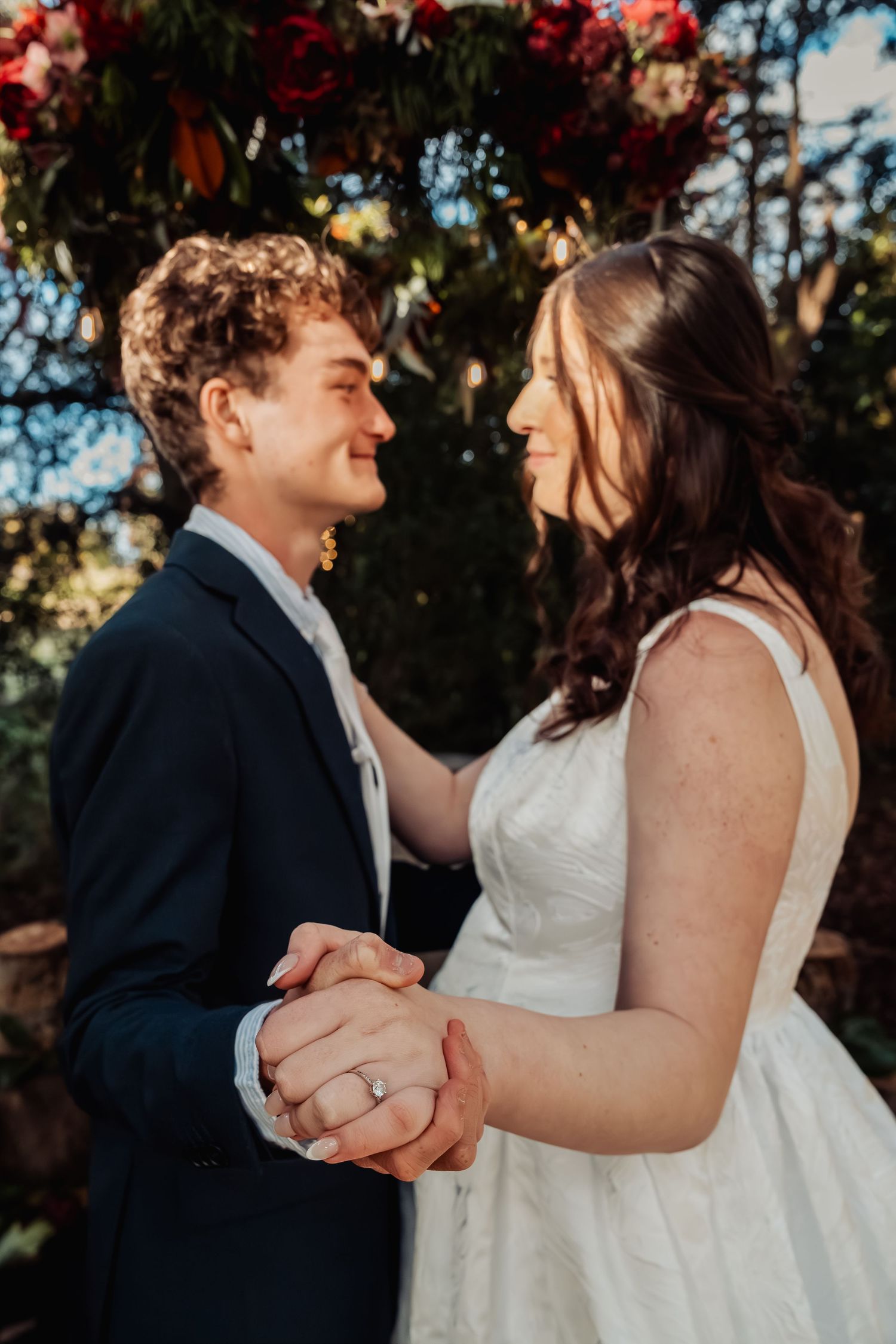 Newlyweds hold hands and gaze lovingly at each other beneath a floral arch of red blooms at their wedding.