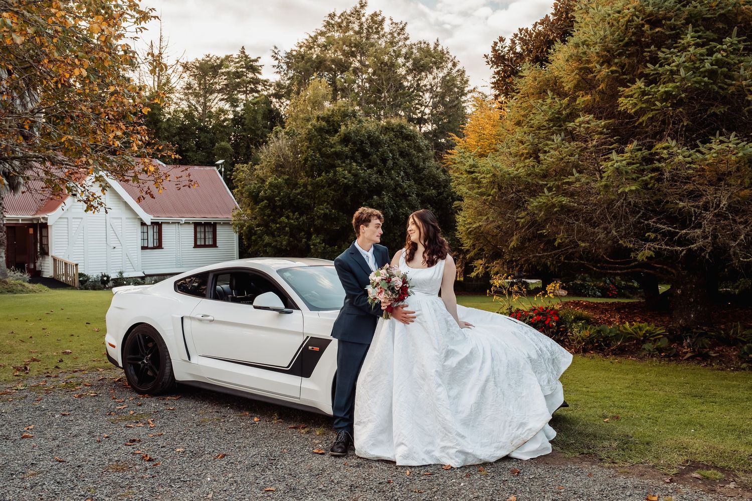 Bride and groom stand together beside a white Ford Mustang on a gravel driveway with autumn trees behind them.