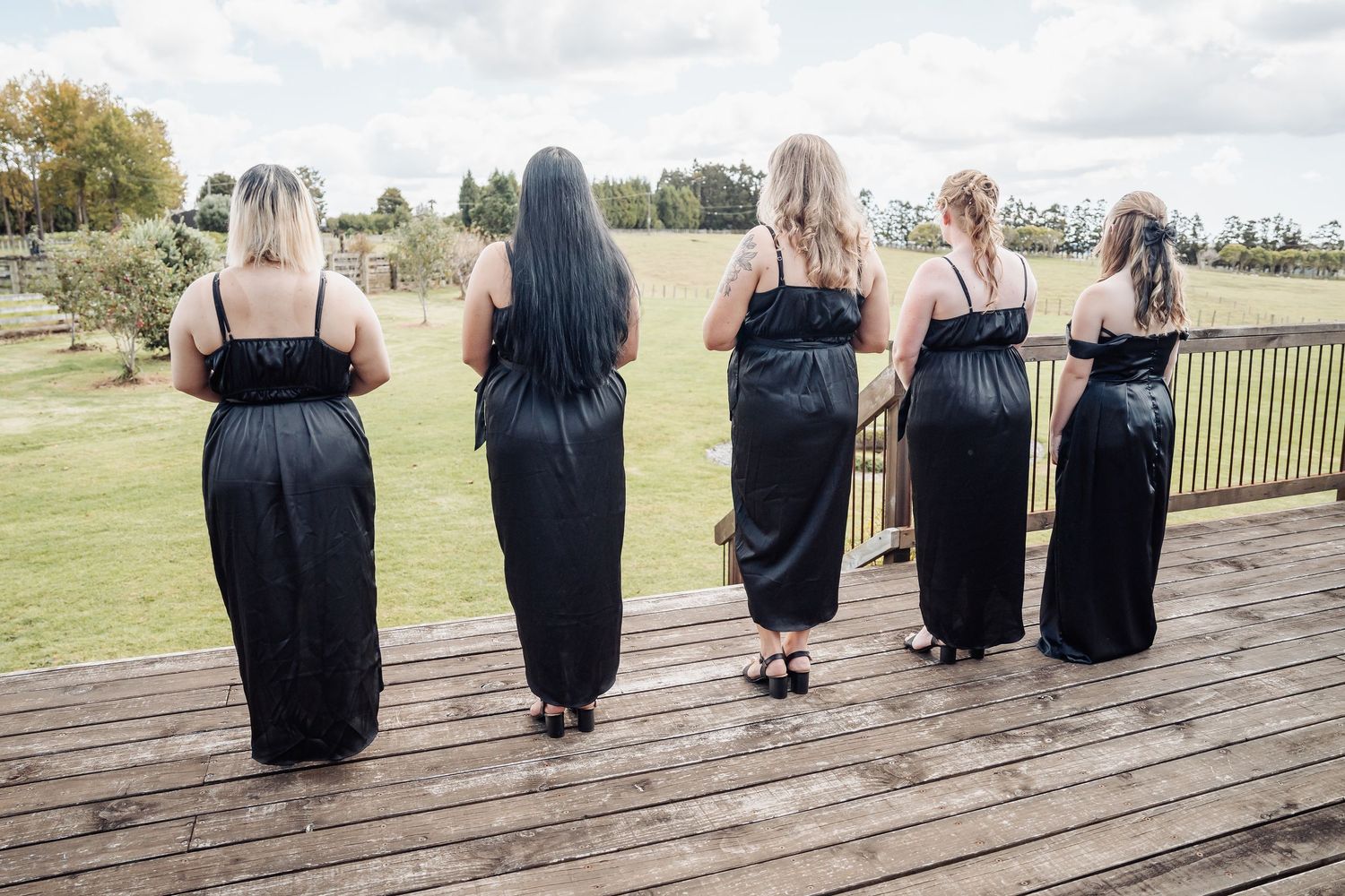 Five women in matching black maxi dresses stand together on a wooden deck overlooking a grassy field.