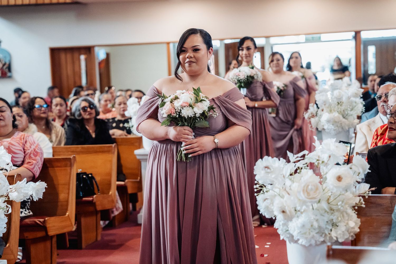 Bridesmaids in mauve dresses holding white floral bouquets during church ceremony.