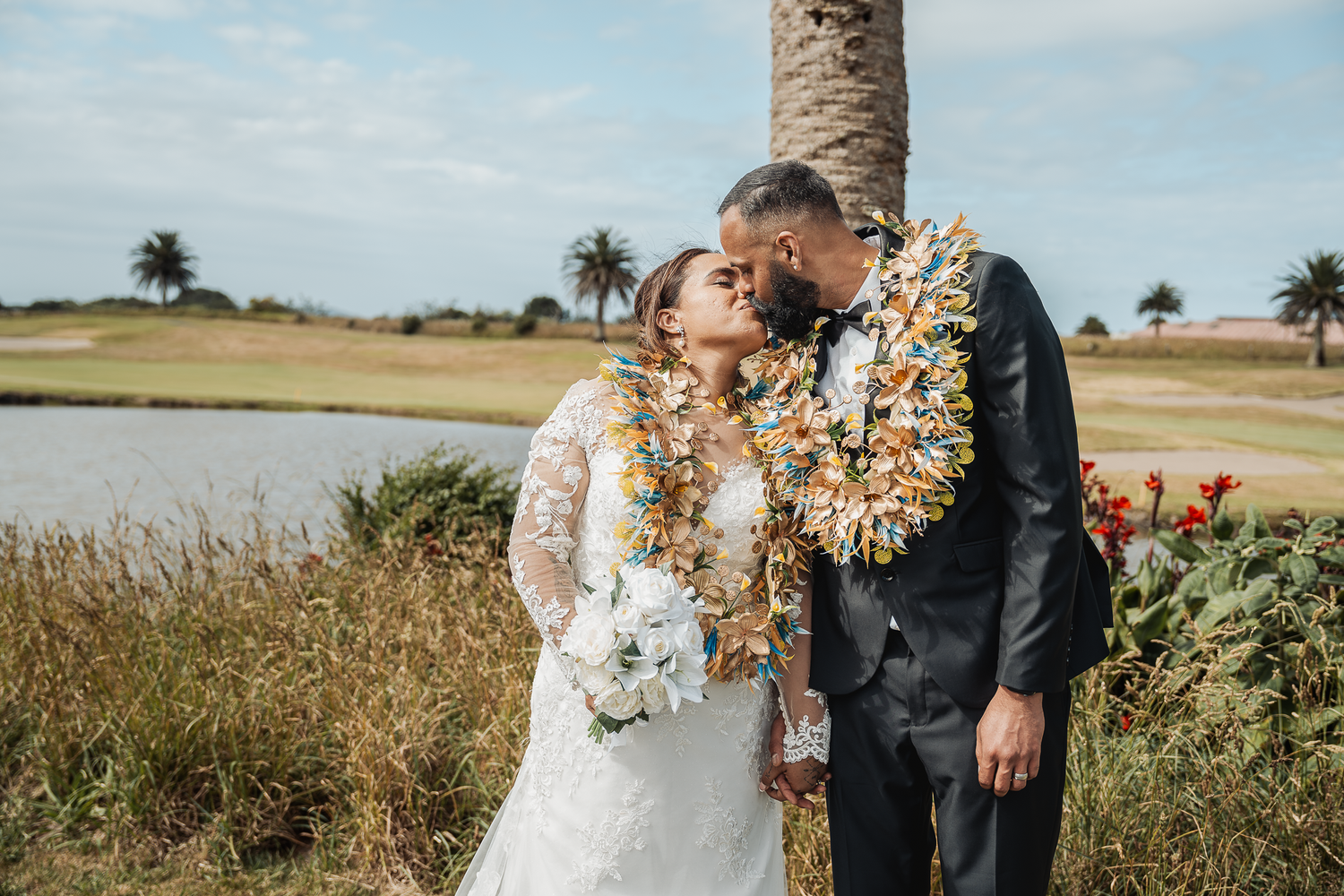 Wedding couple wearing traditional Hawaiian leis embraces in outdoor setting.