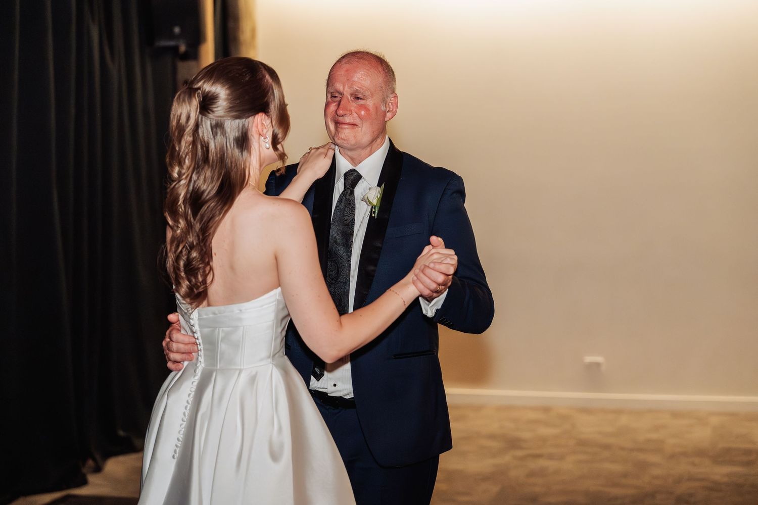 Bride and older man share a tender father-daughter dance on a dimly lit wedding reception dance floor.