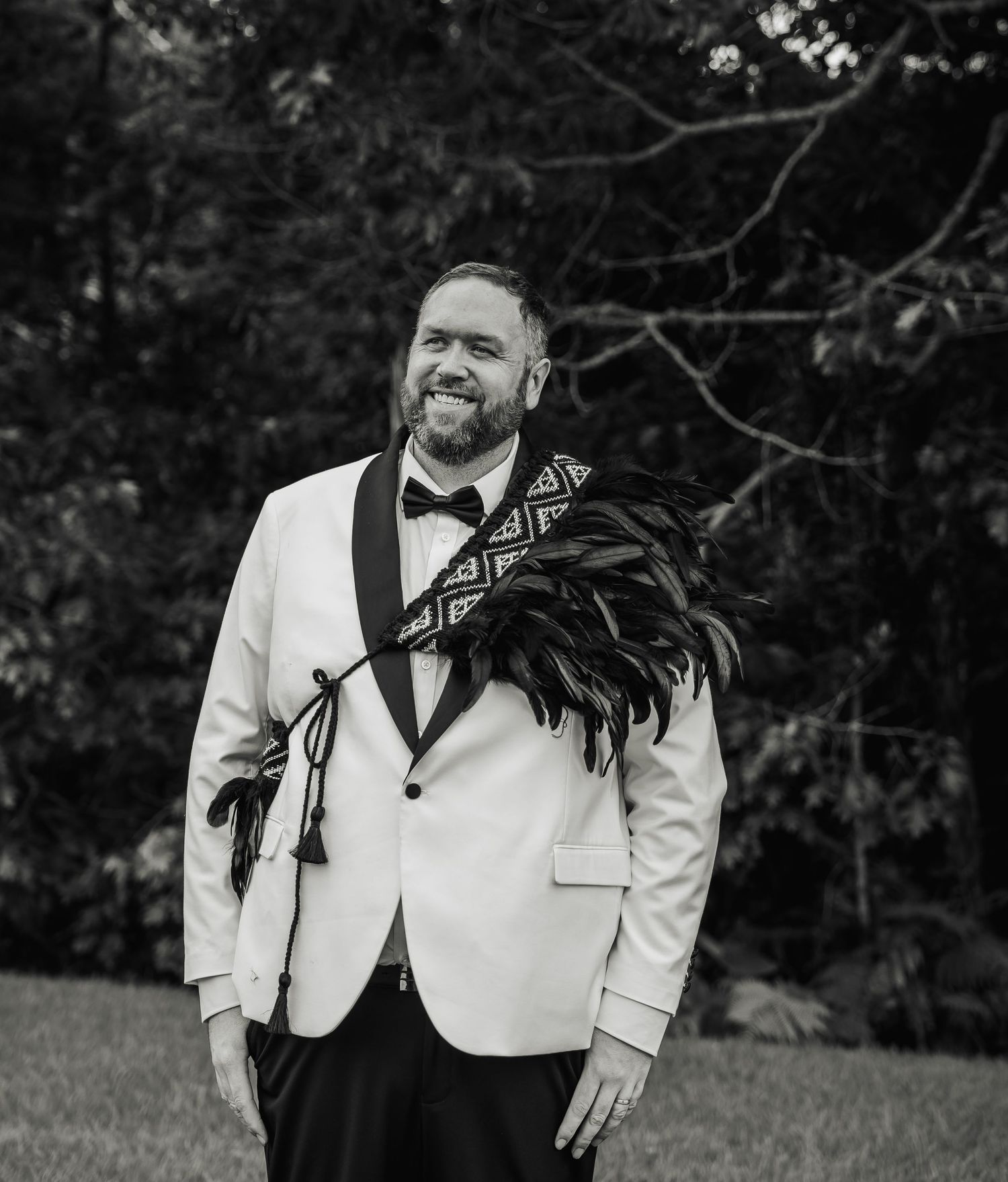 Groom in white tuxedo with traditional Maori feather cloak smiles outdoors in black and white photo.