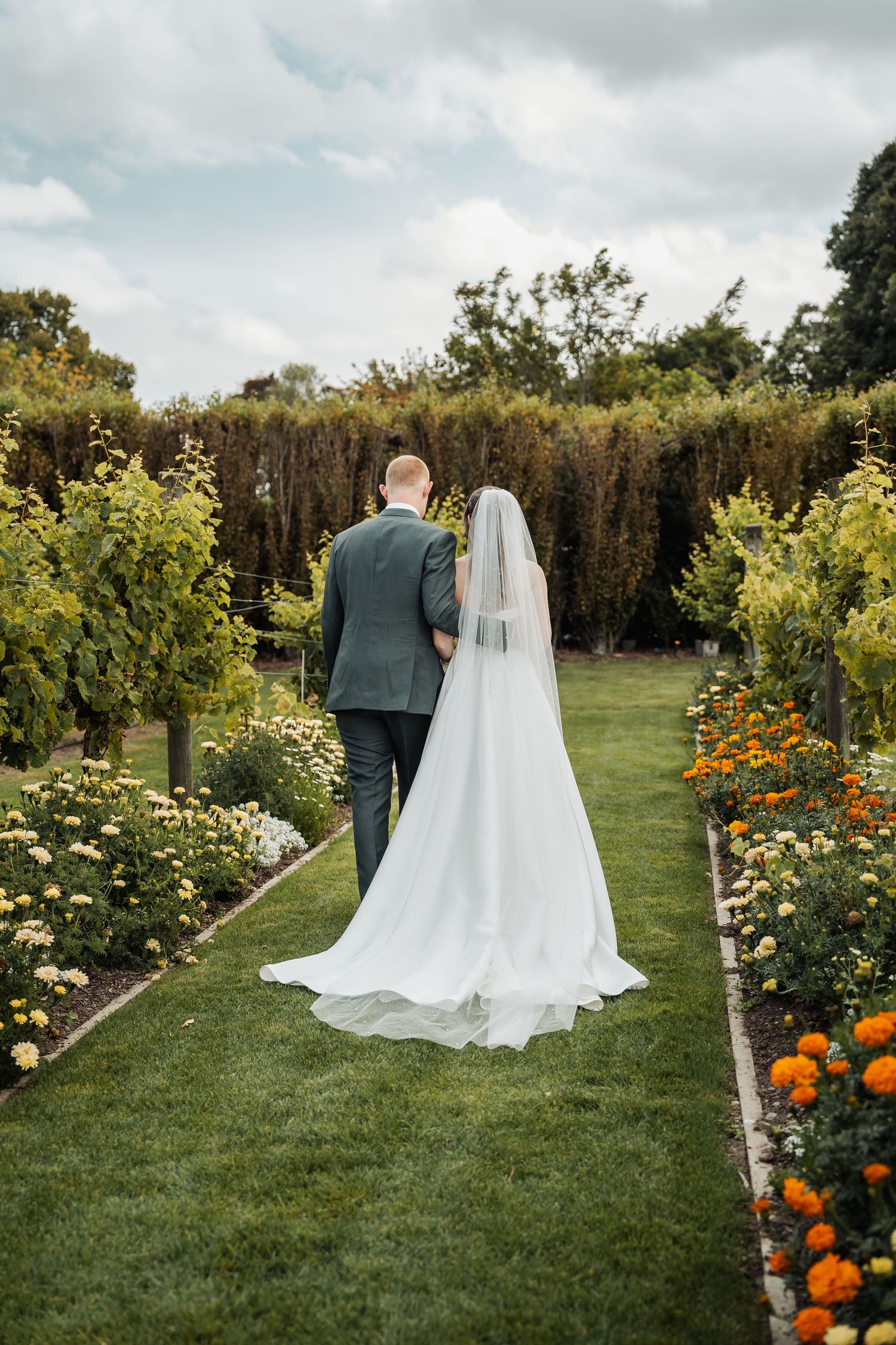Bride and groom walk hand in hand through a lush garden path lined with colorful flowers and green hedges.