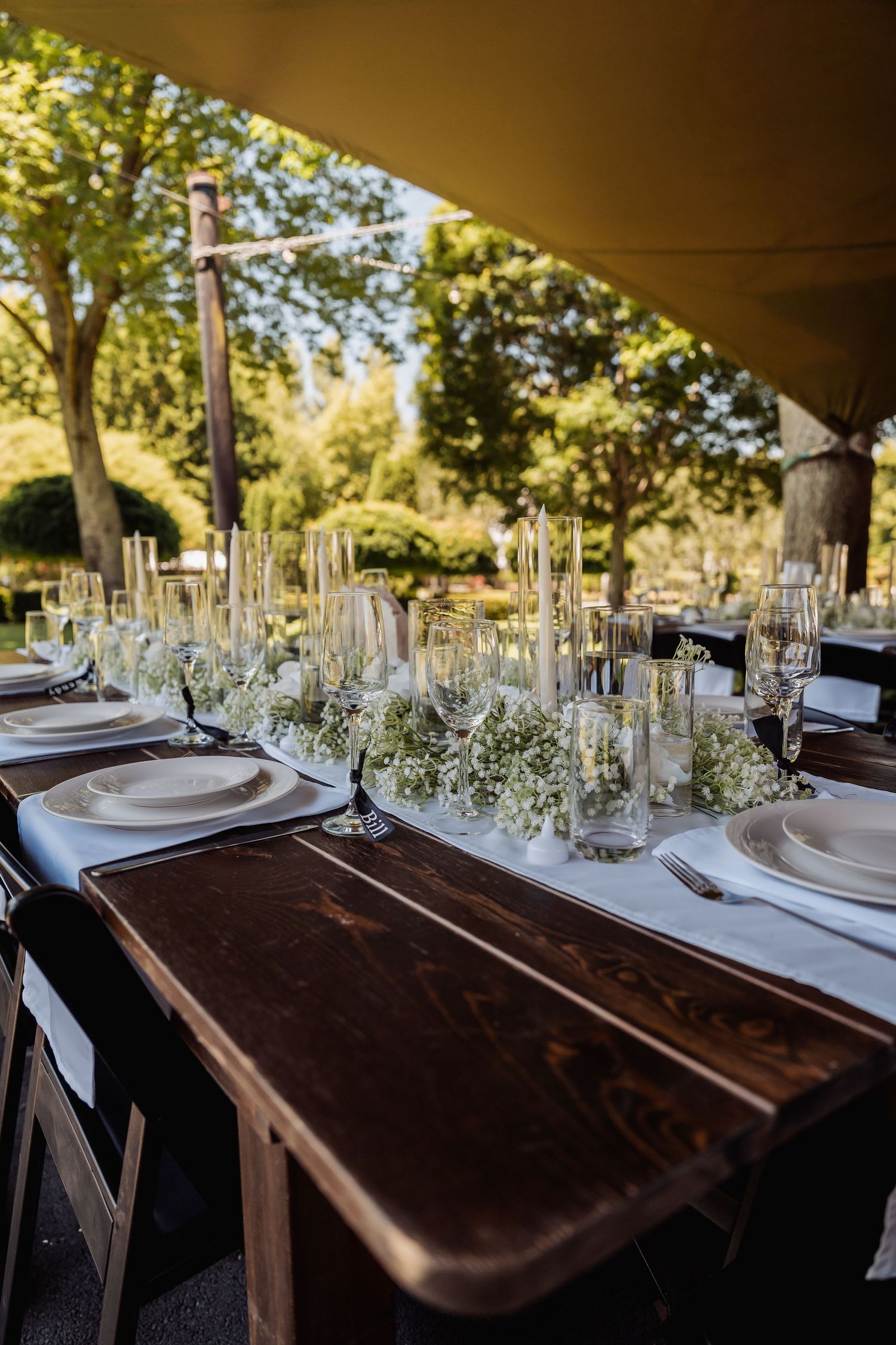 Elegant outdoor wedding reception table with white floral centerpieces, tall candles, and glassware on a rustic wood table.