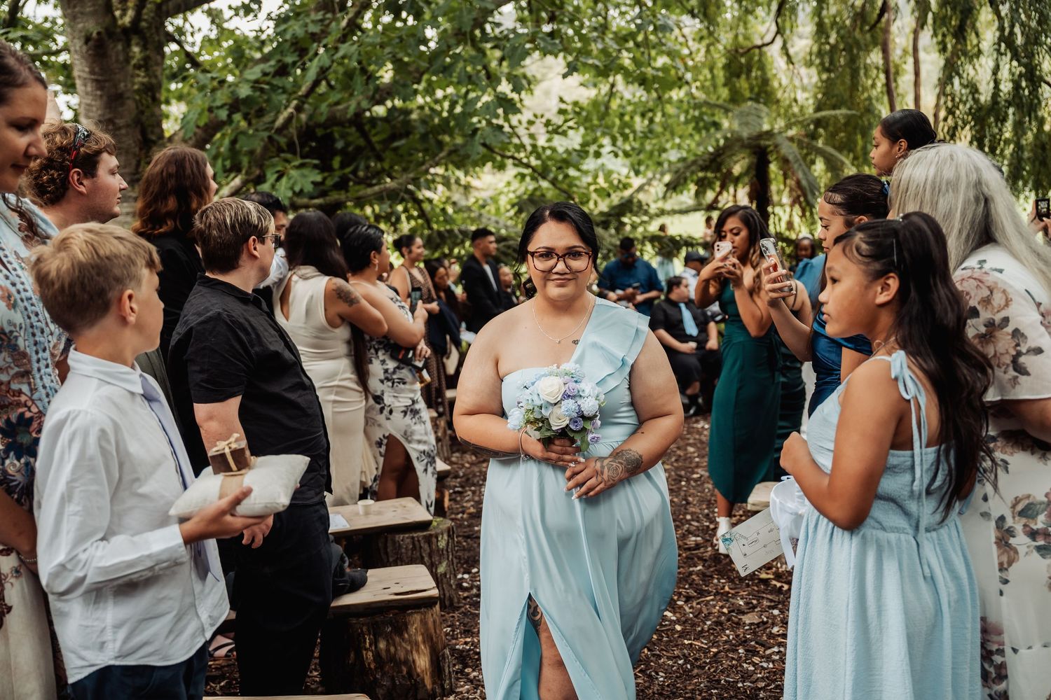 Bridesmaid in light blue dress walks down outdoor woodland wedding aisle among smiling guests.