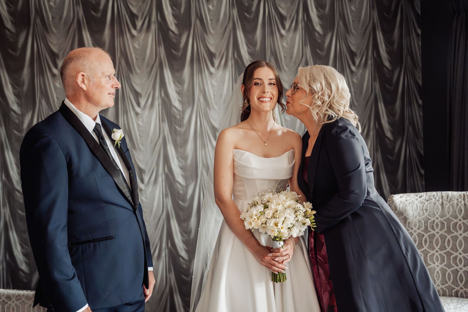 Bride in white gown holds bouquet while older woman kisses her cheek, man in navy suit stands nearby.