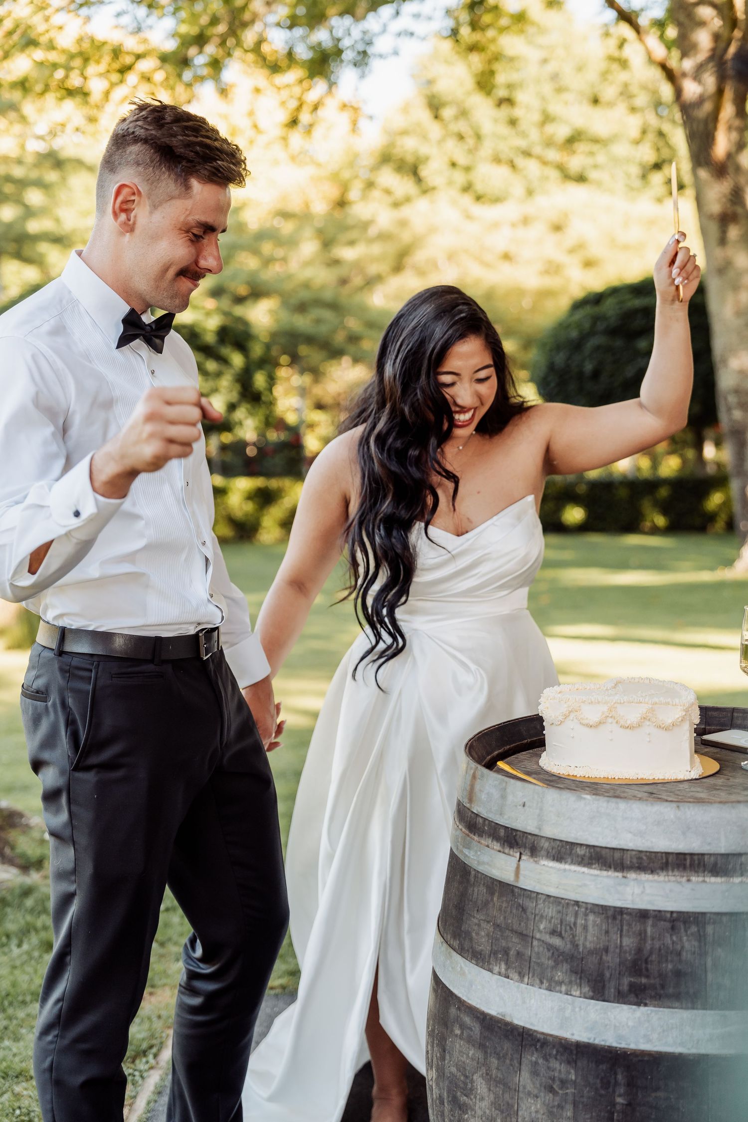 Bride and groom joyfully cutting their wedding cake on a wine barrel outdoors in a lush green garden.