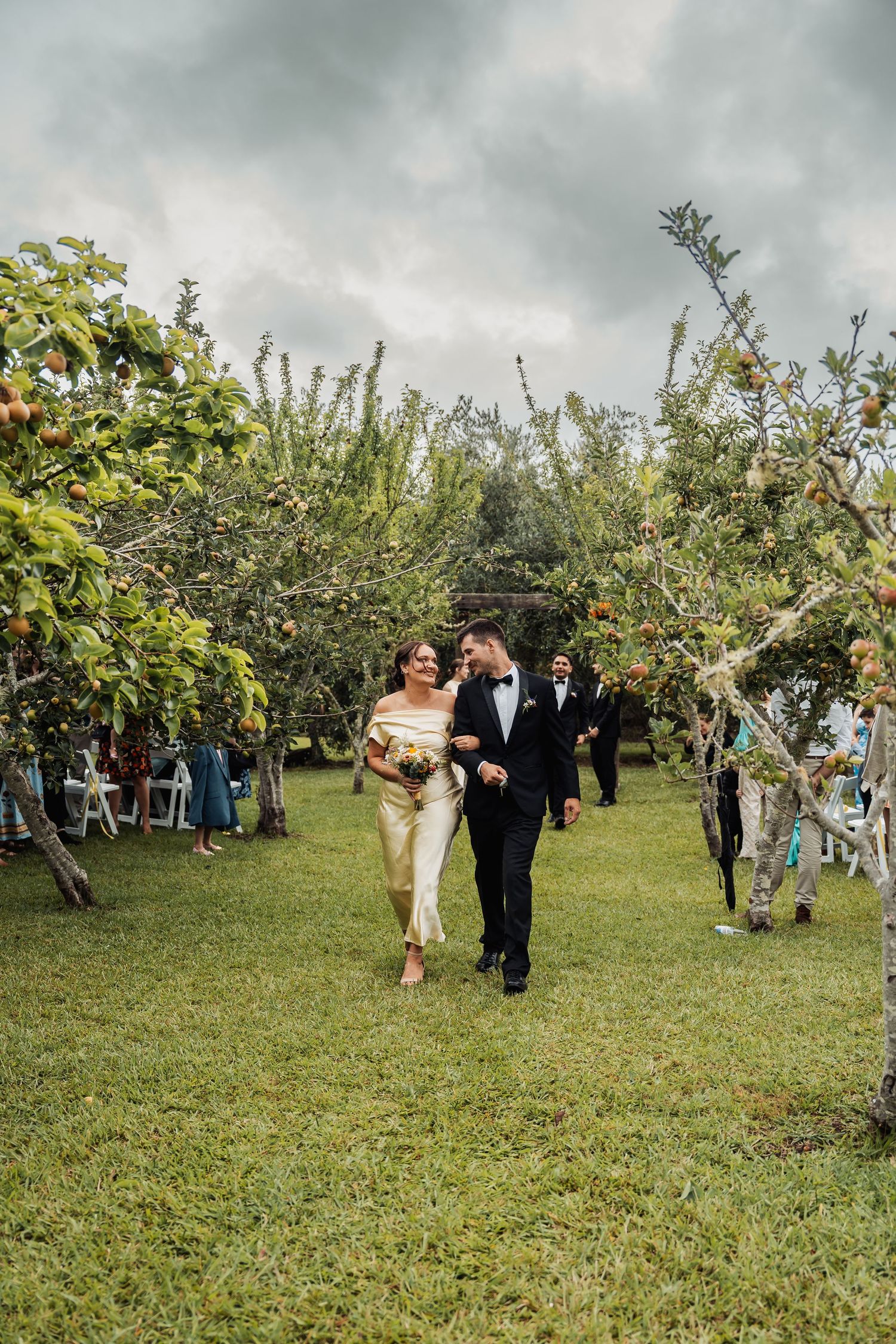 Bride and groom walk hand-in-hand through a lush garden orchard aisle during their outdoor wedding ceremony.