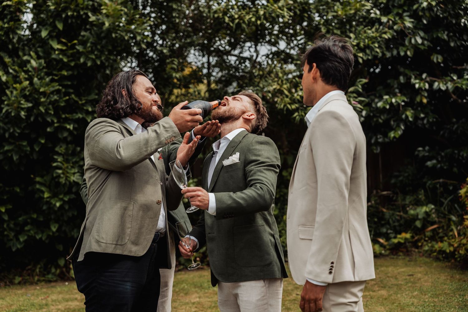 Groomsmen celebrating outdoors, one pouring a drink into another's mouth at a garden wedding.
