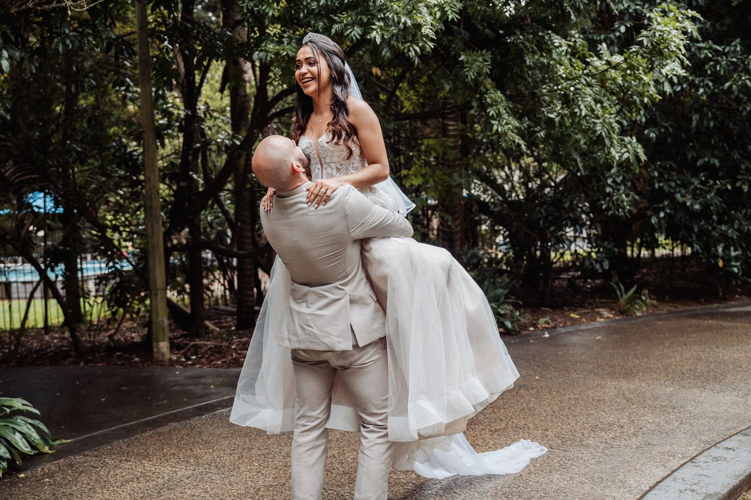 Groom lifts smiling bride in white gown outdoors surrounded by lush green trees after wedding ceremony.