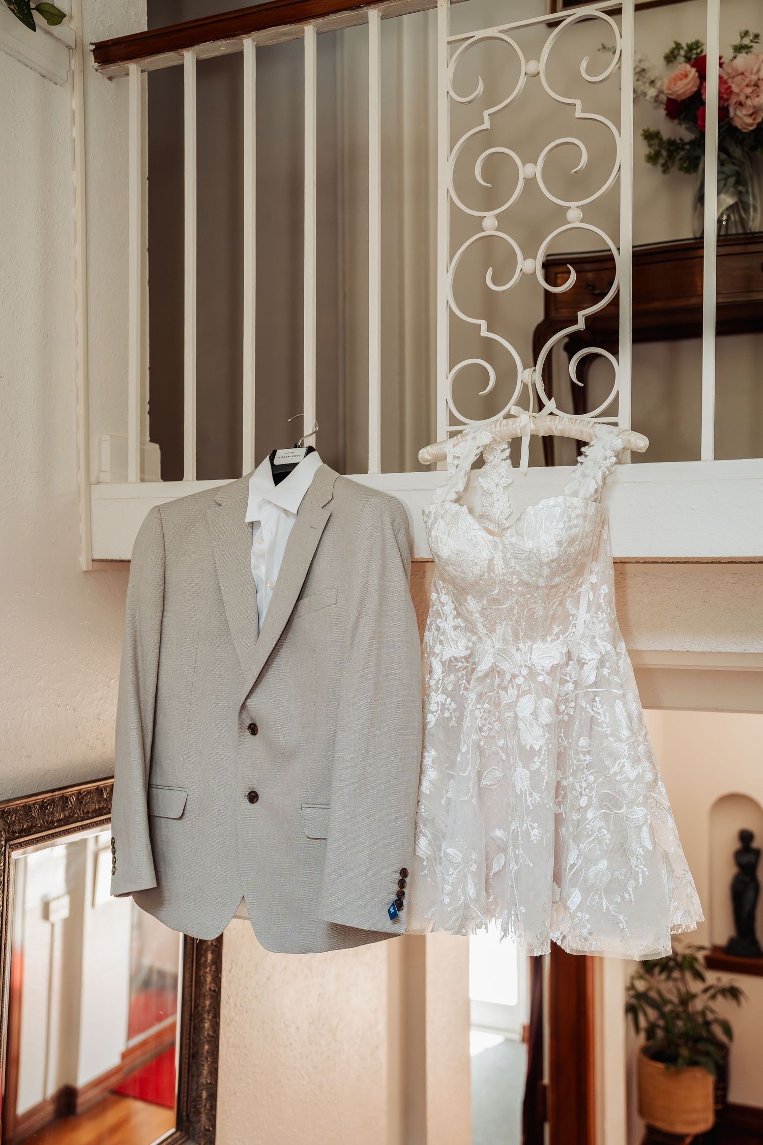 Gray suit and white lace wedding dress hanging on ornate white staircase railing in elegant home.