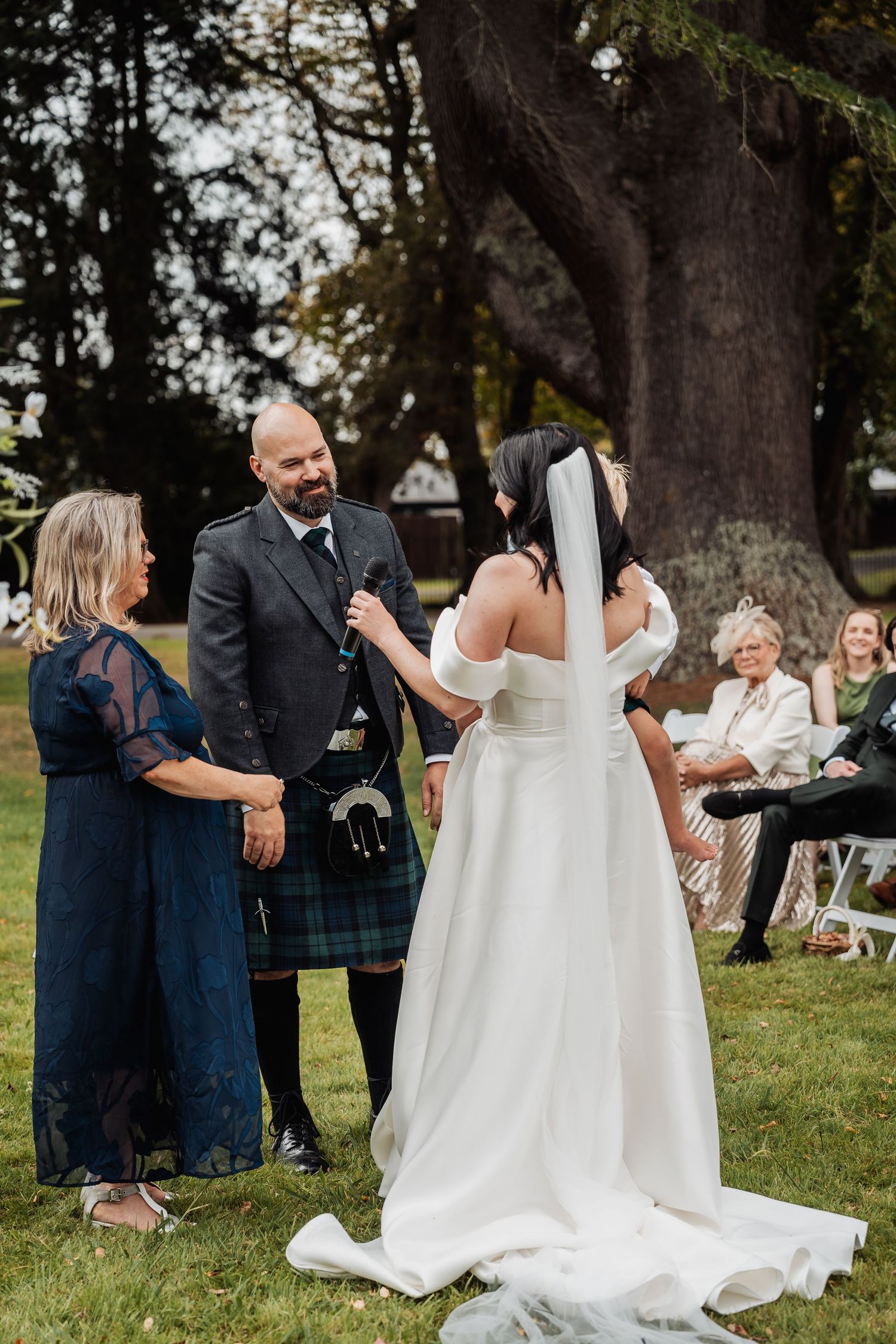 Bride in white off-shoulder gown exchanges vows with groom in Scottish tartan kilt at outdoor wedding ceremony.