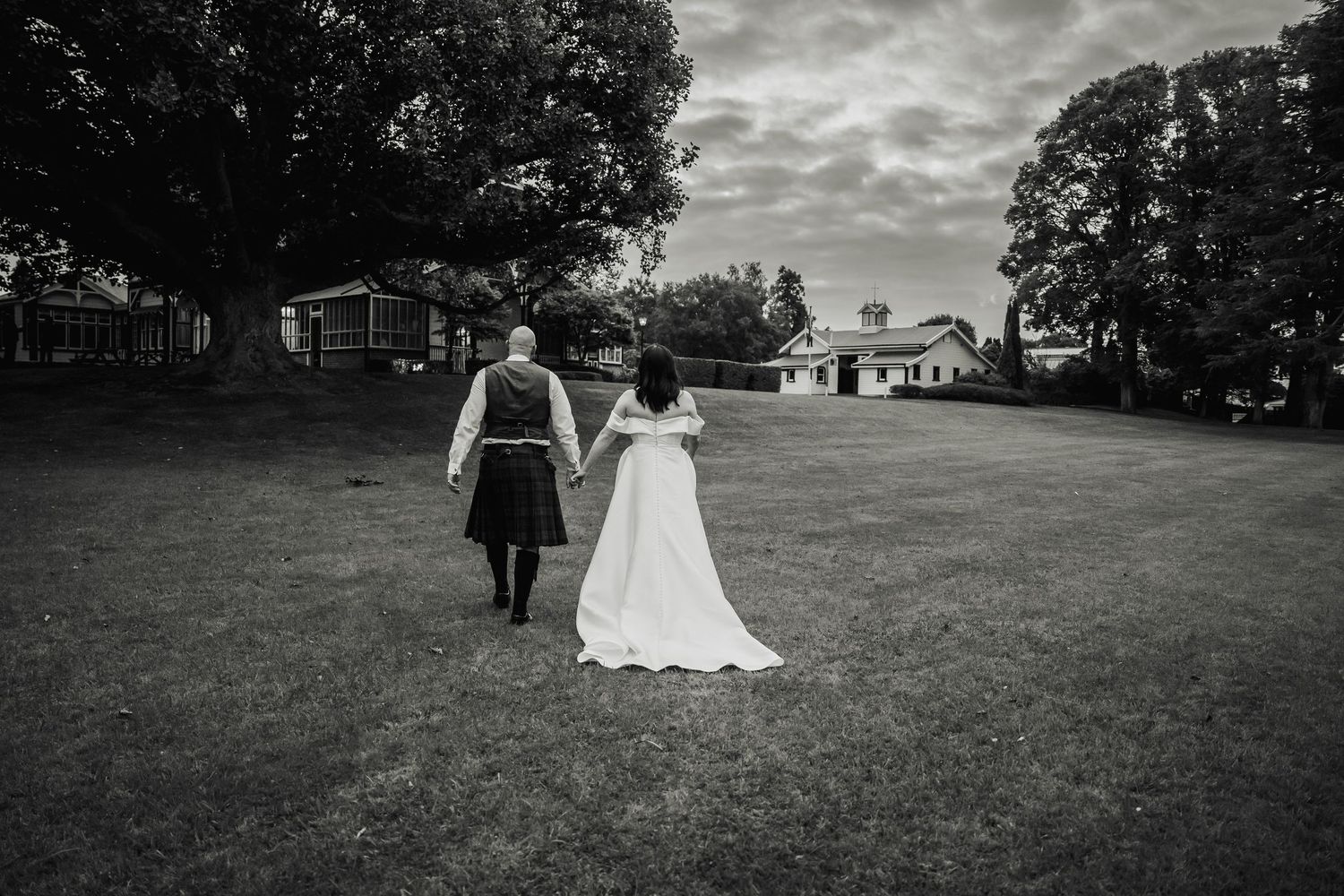 Black and white photo of a bride and groom holding hands walking across a green lawn toward rustic buildings.