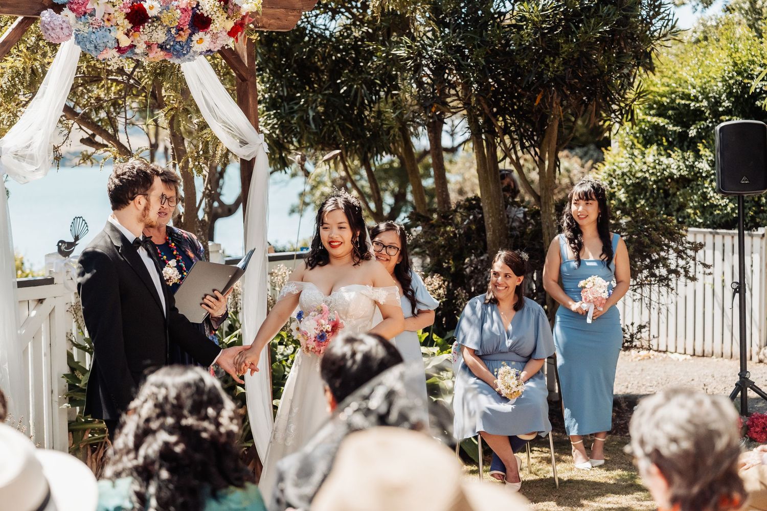 Couple exchanging vows at outdoor wedding ceremony with bridesmaids in blue dresses and floral arch.