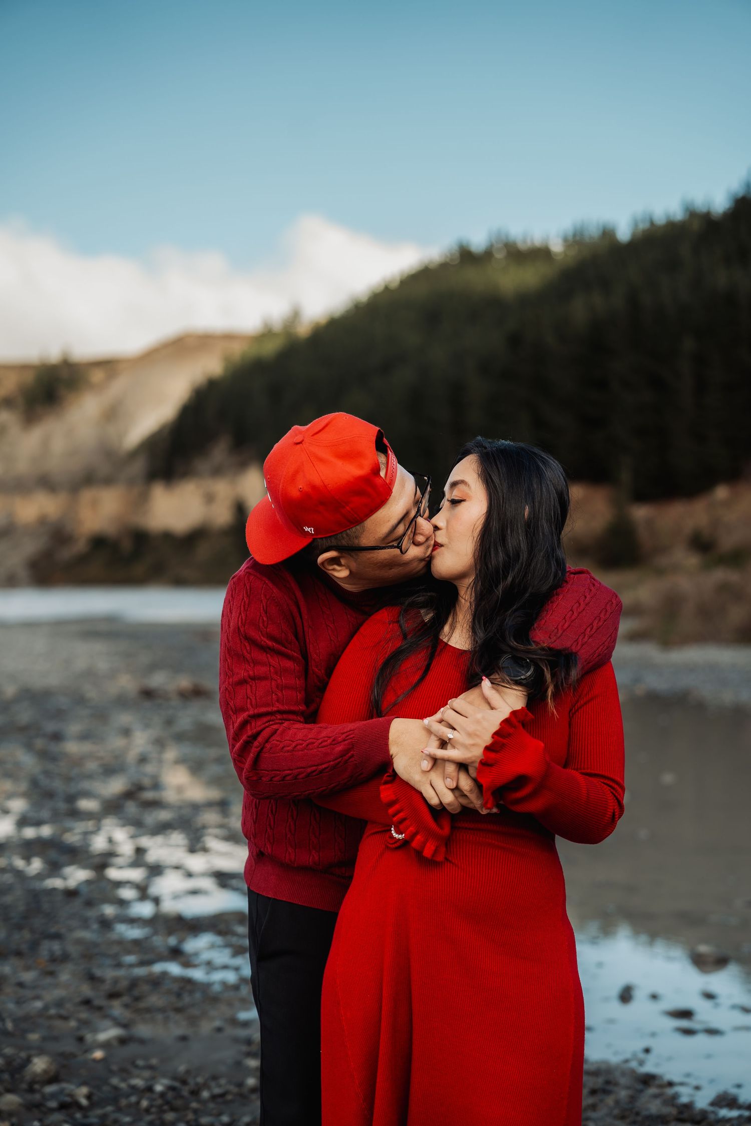 Couple sharing a romantic kiss by a rocky riverbank with a green mountain backdrop on a sunny day.