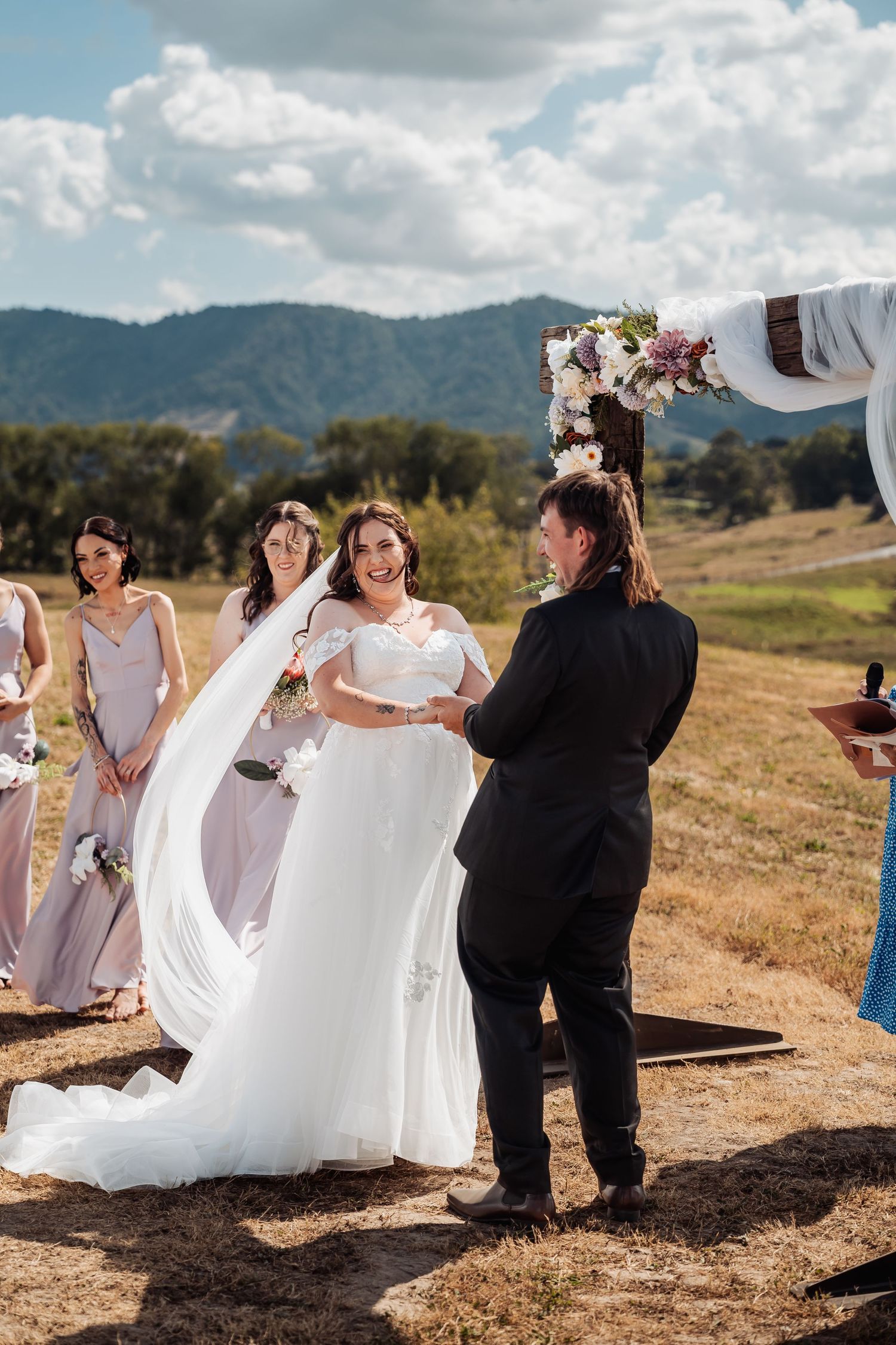 Bride in white gown exchanges vows with partner at outdoor hilltop ceremony with mountain backdrop.