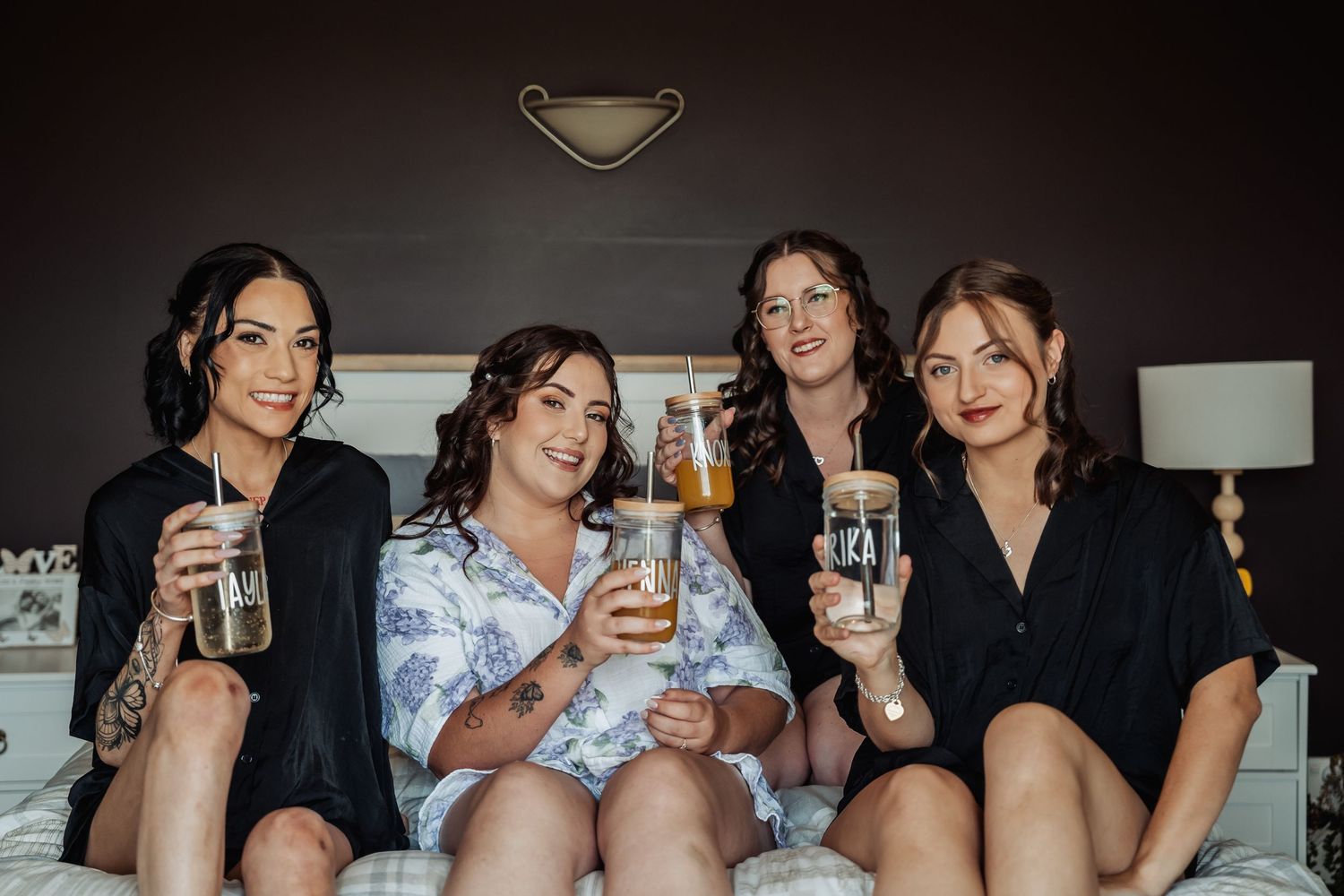 Four bridesmaids in robes sitting on a bed smiling and holding iced drinks during wedding morning preparations.