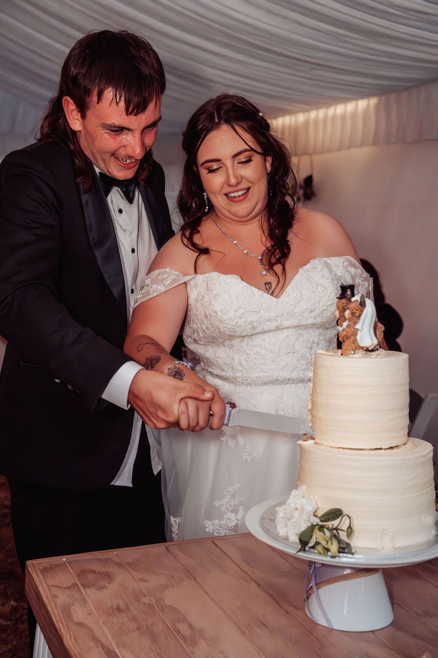 Smiling bride and groom cut three-tier white wedding cake together inside decorated reception marquee.