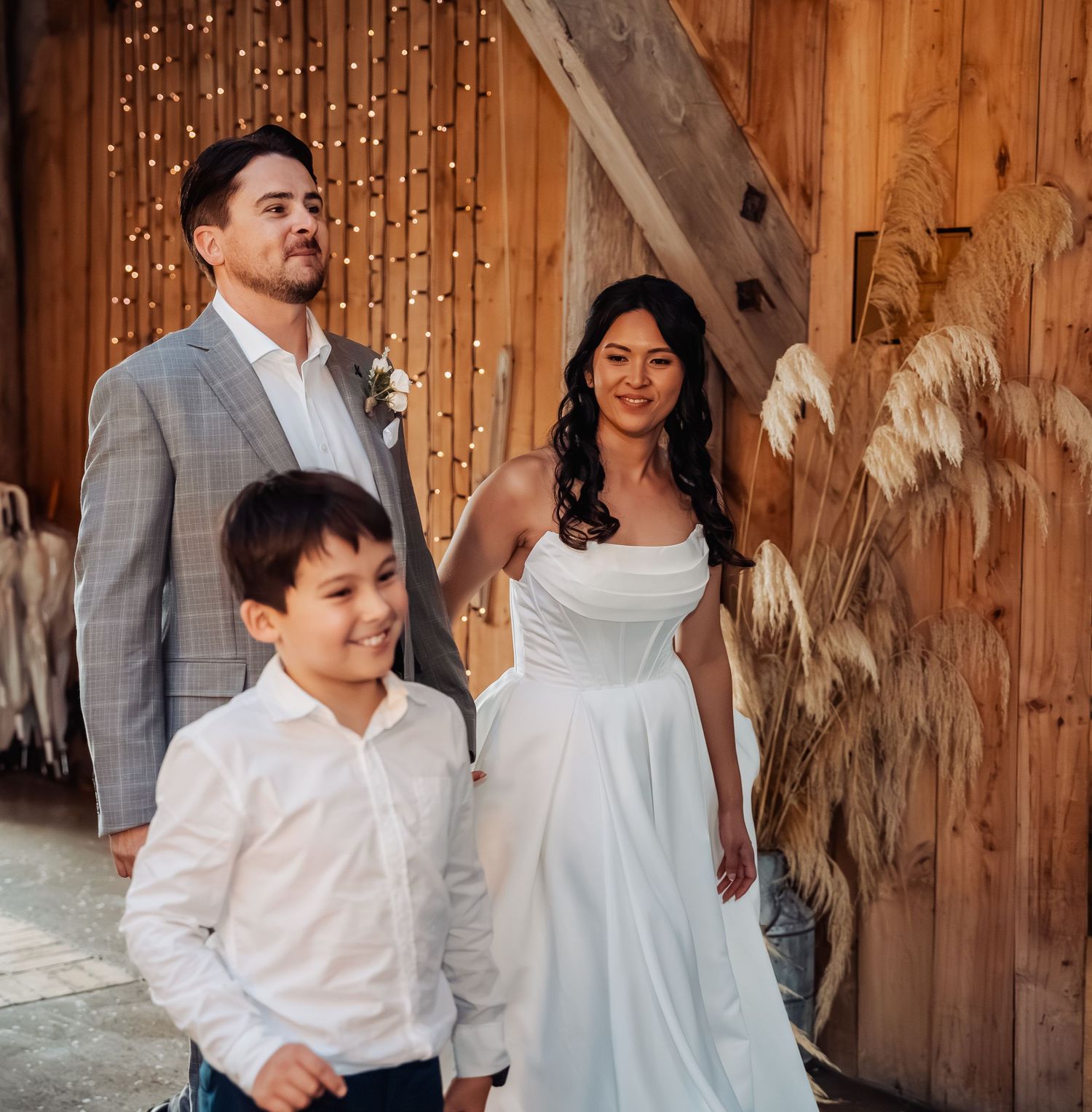 Bride in white gown and groom in gray suit stand with young boy in barn wedding venue with fairy lights.