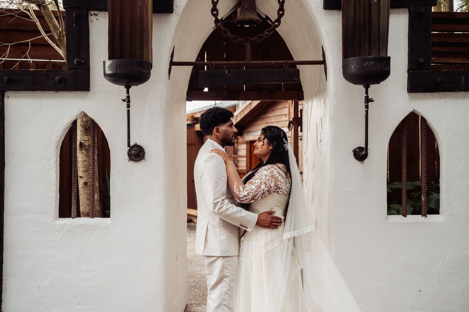 Bride and groom share romantic embrace at entrance of white chapel with ornate dark lanterns.