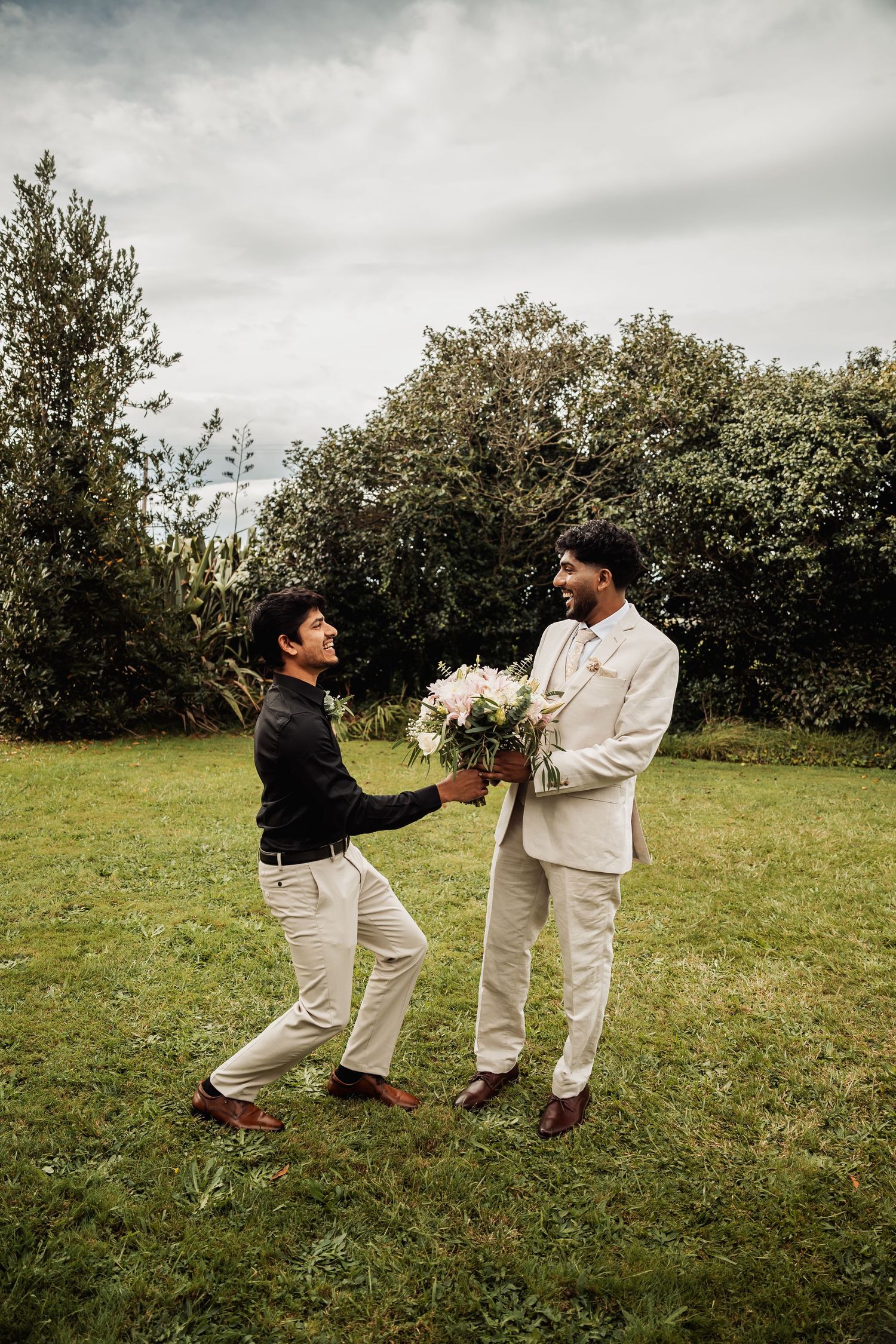 Groom in beige suit receiving bouquet from groomsman in black on green lawn.