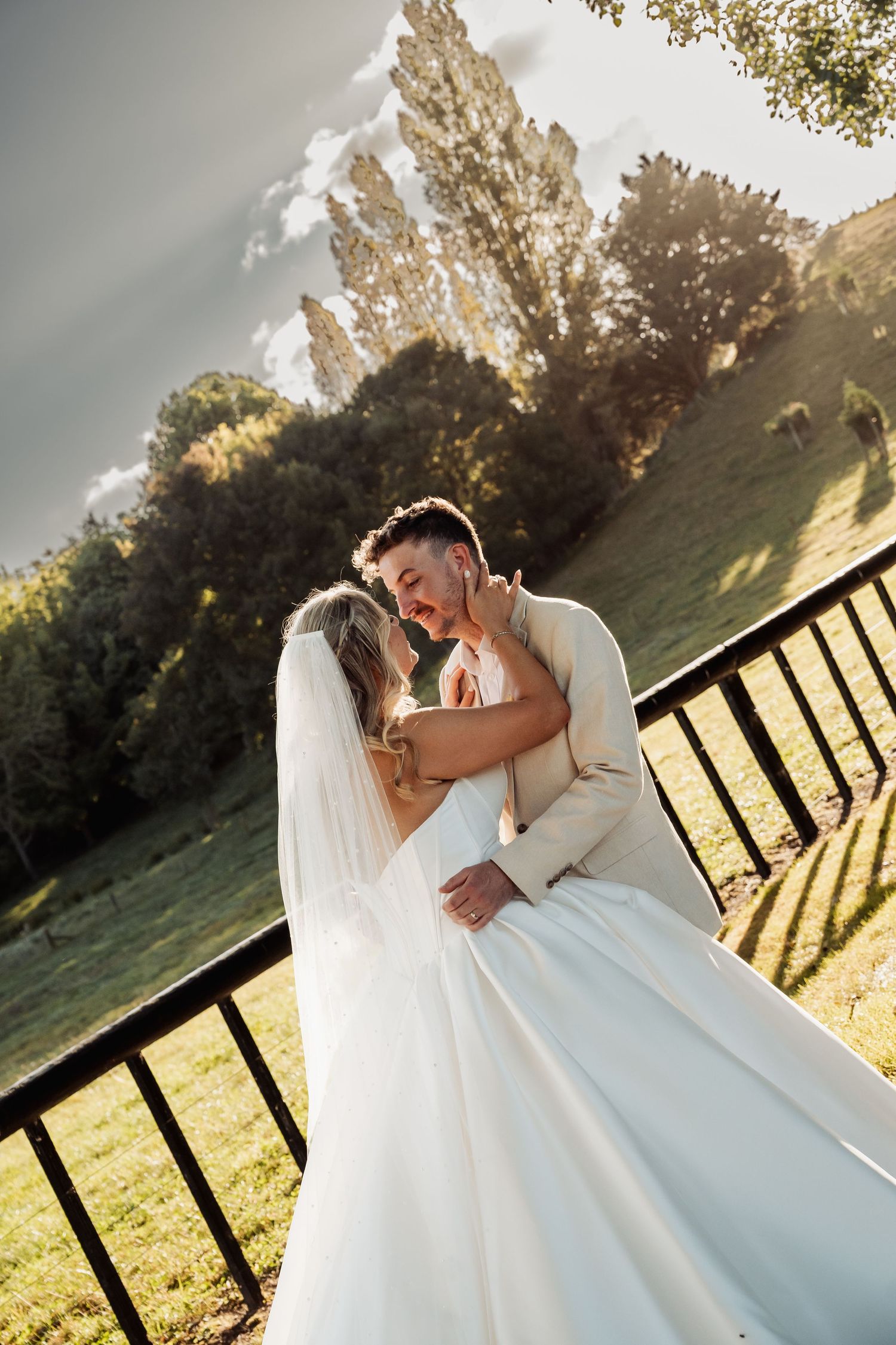 Bride and groom embracing romantically near a wooden fence with lush green hills behind them.