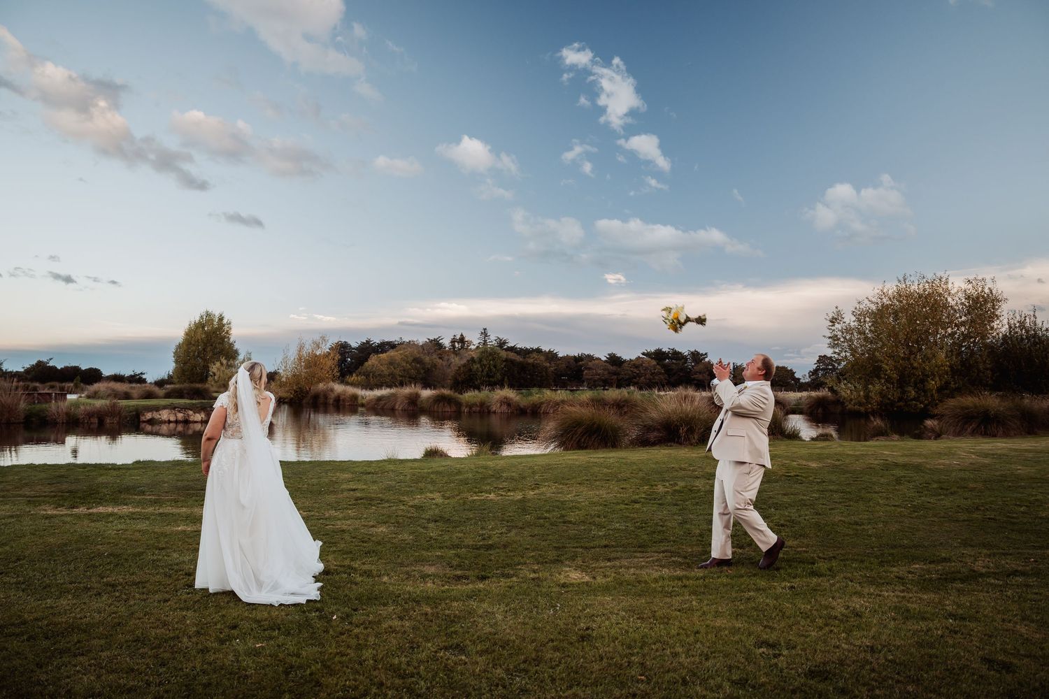 Bride and groom tossing bouquet by scenic lakeside at sunset during outdoor wedding celebration.