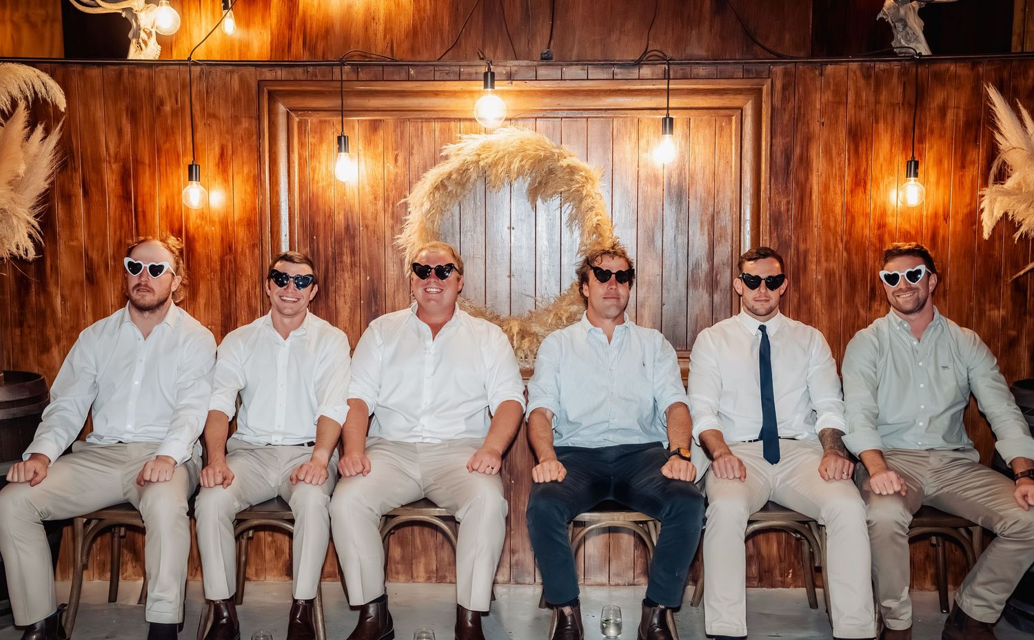 Six groomsmen in white shirts and khaki pants sit together wearing sunglasses inside a rustic barn venue.