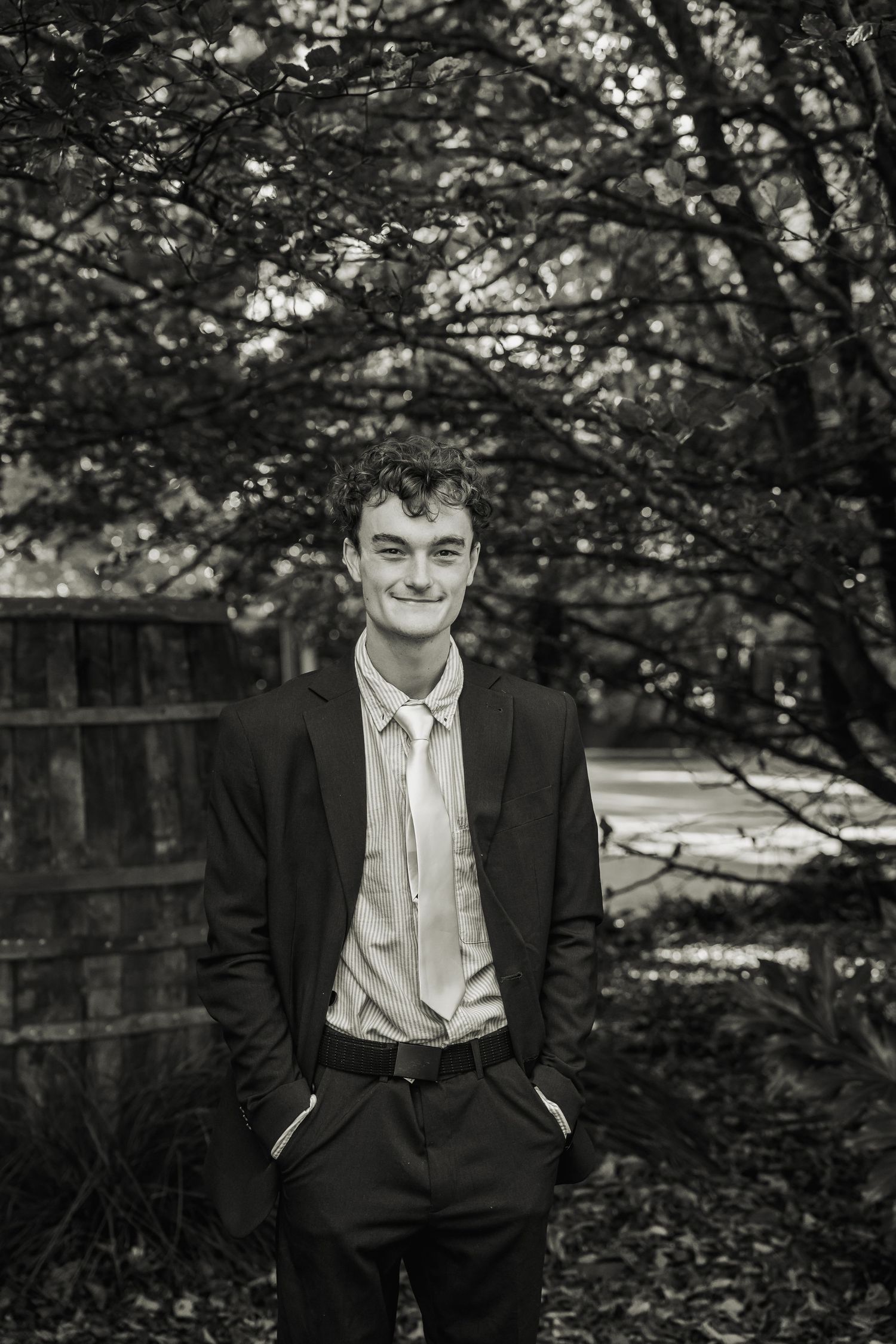 Young man in a black suit smiling outdoors under trees in a black and white portrait photo.