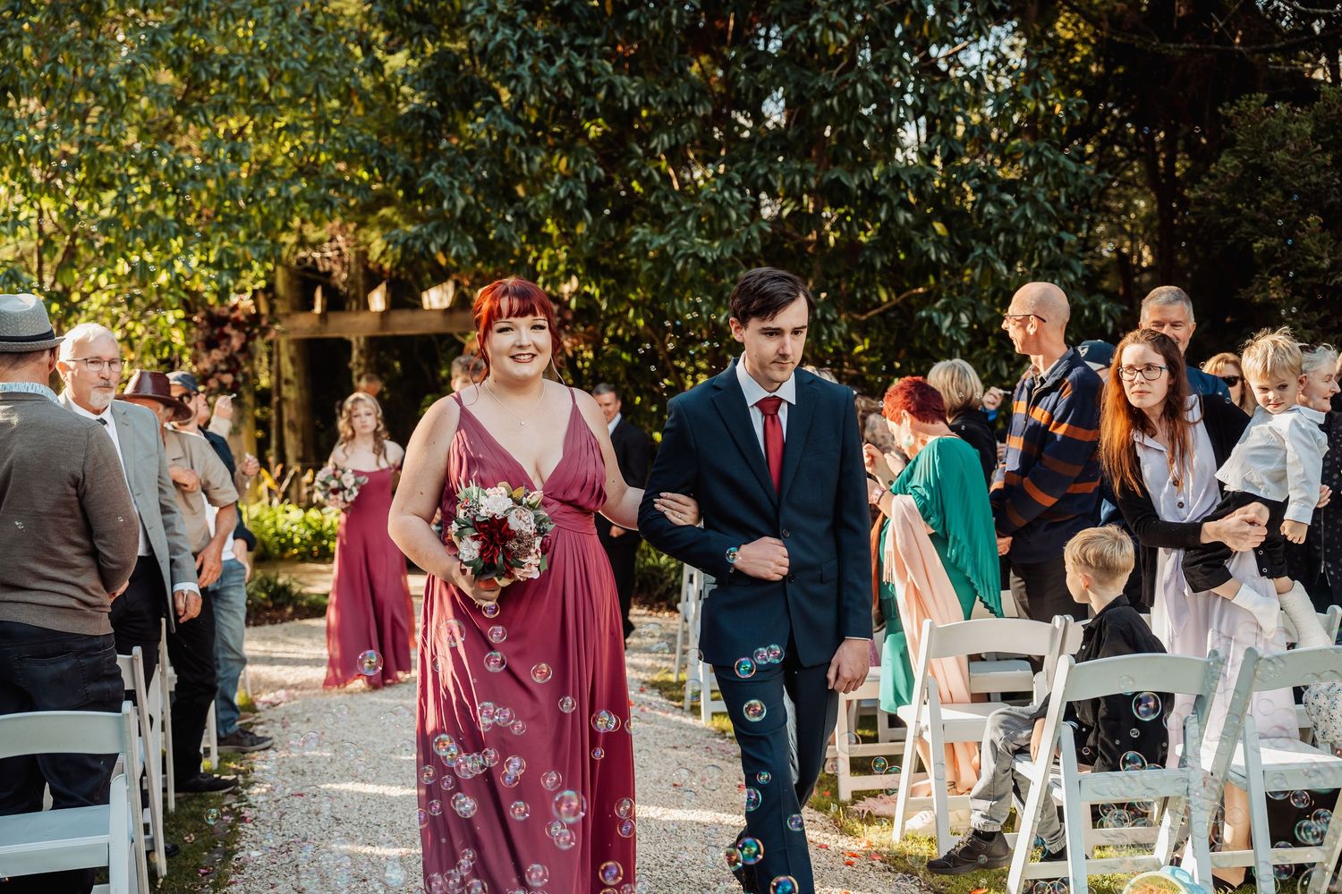 Bridesmaid in pink floral dress walks down outdoor wedding aisle with groomsman in navy suit.
