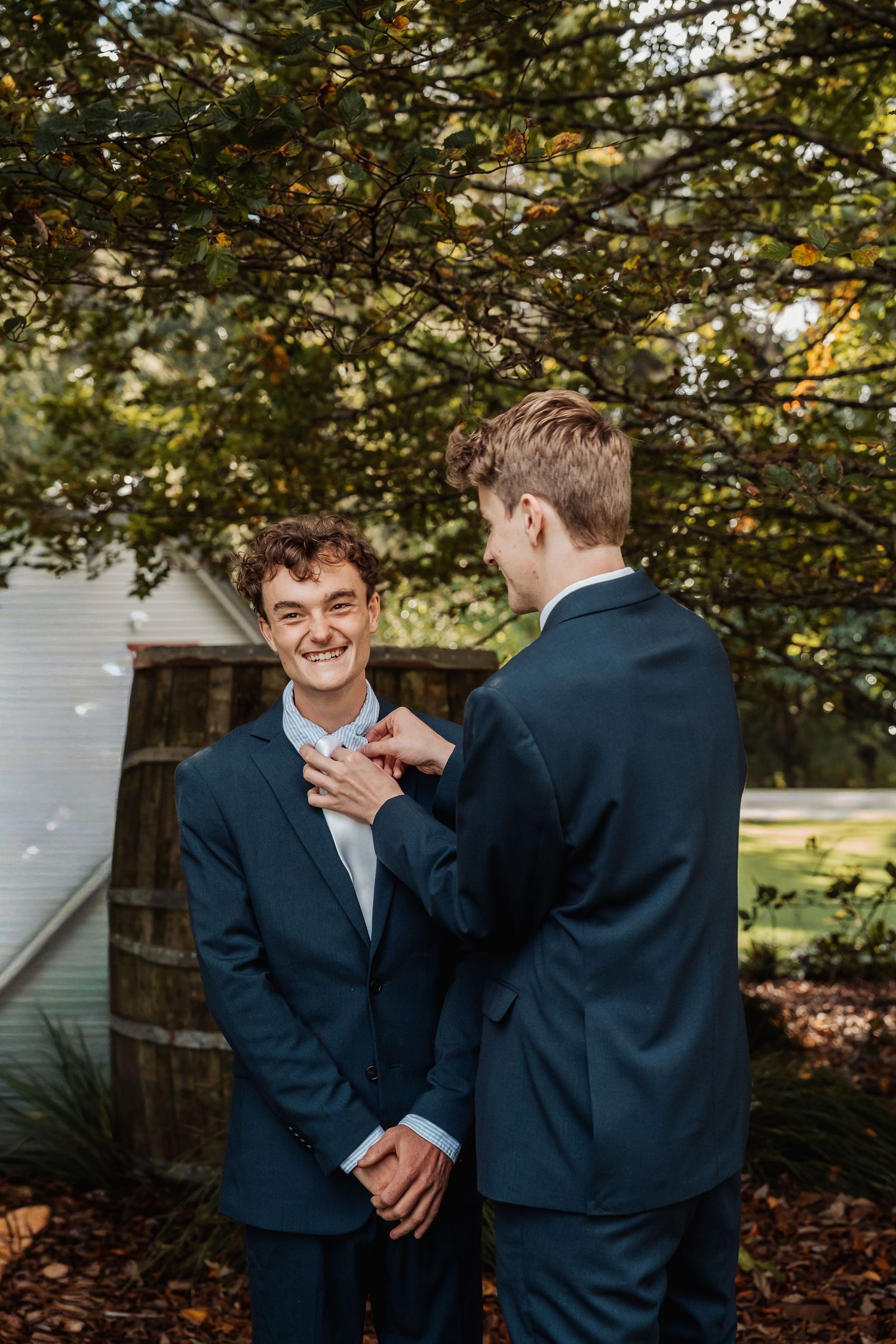 Two groomsmen in navy suits adjusting boutonniere outdoors among green trees.