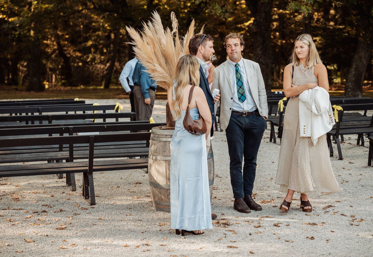 Wedding guests in elegant attire socializing outdoors among decorated benches at a rustic venue.