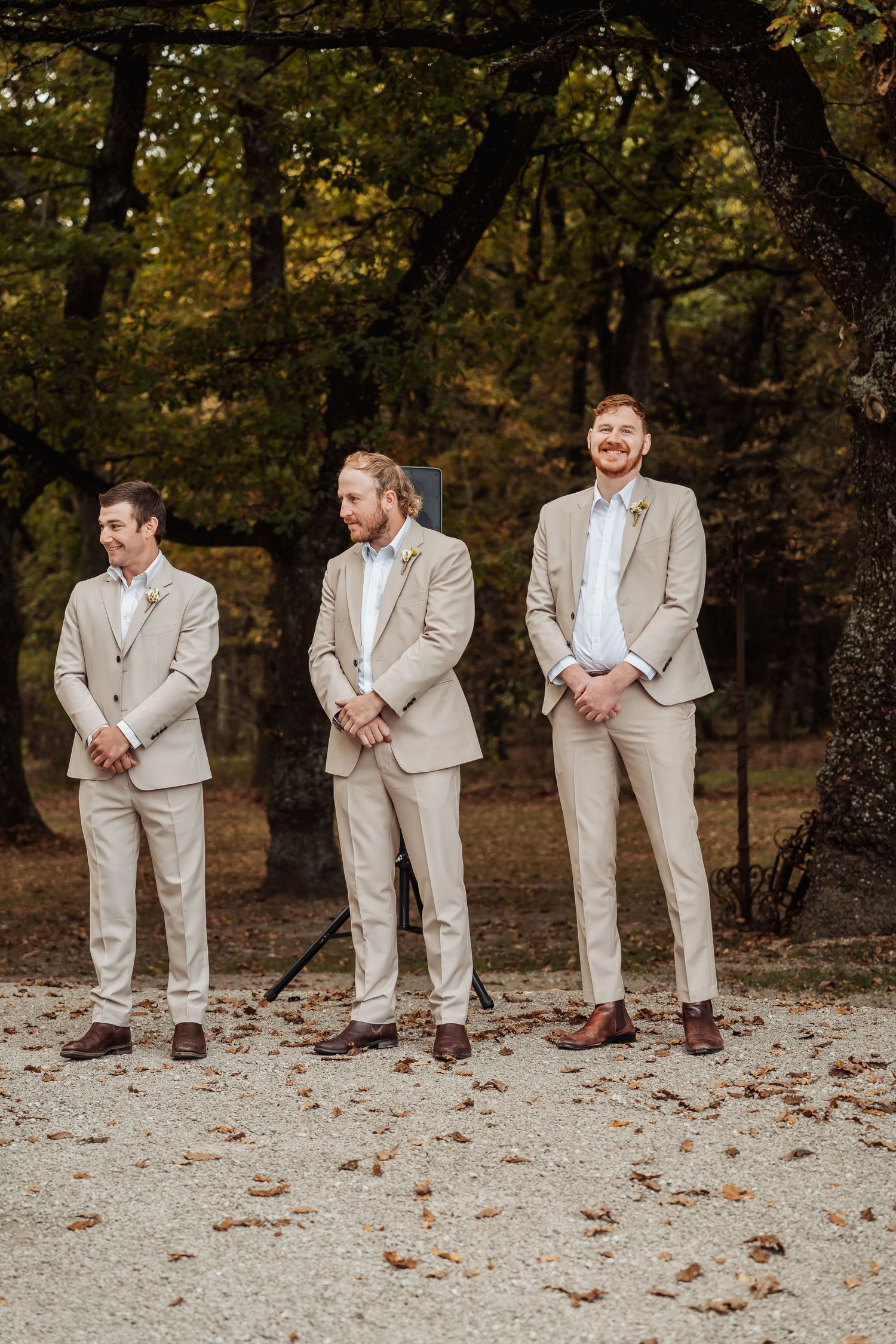 Three groomsmen in matching tan suits stand outdoors among autumn trees at a wedding ceremony.