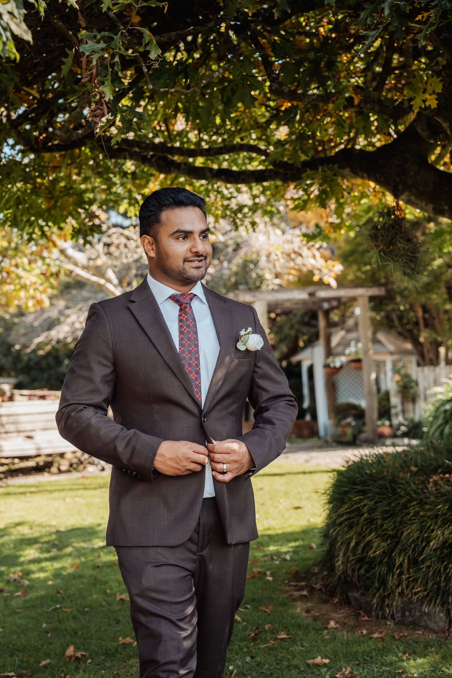 Groom in brown suit with floral boutonniere standing outdoors under tree at garden wedding venue.